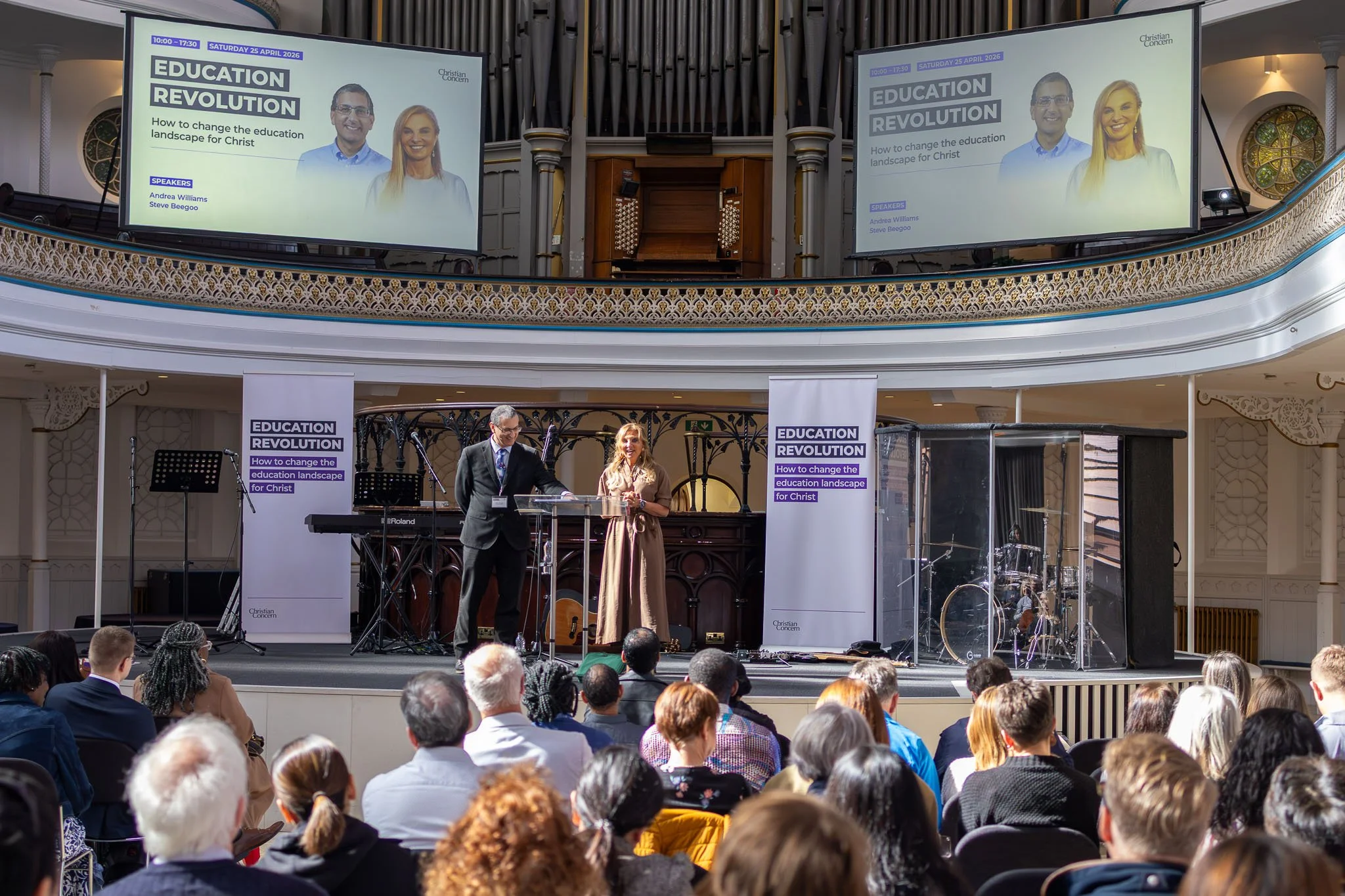 A large audience gathered in a church, attending a presentation or event titled 'Education Revolution' with the subtitle 'How to change the education landscape for Christ.' On stage, a man in a suit and a woman in a dress are standing behind a clear 