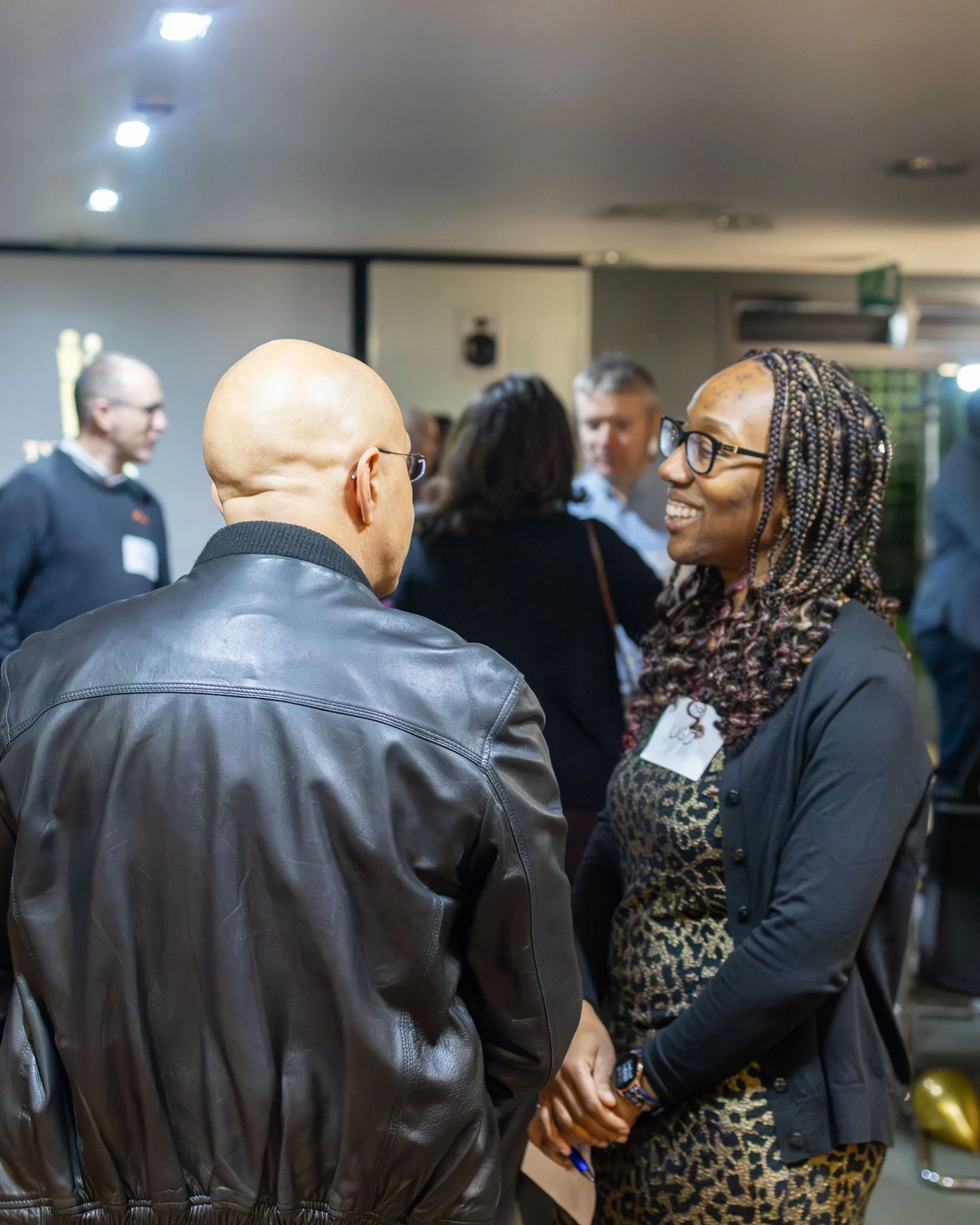 People engaging in conversation at an indoor event, with a woman smiling and wearing glasses, and a man with a shaved head and glasses in the foreground.