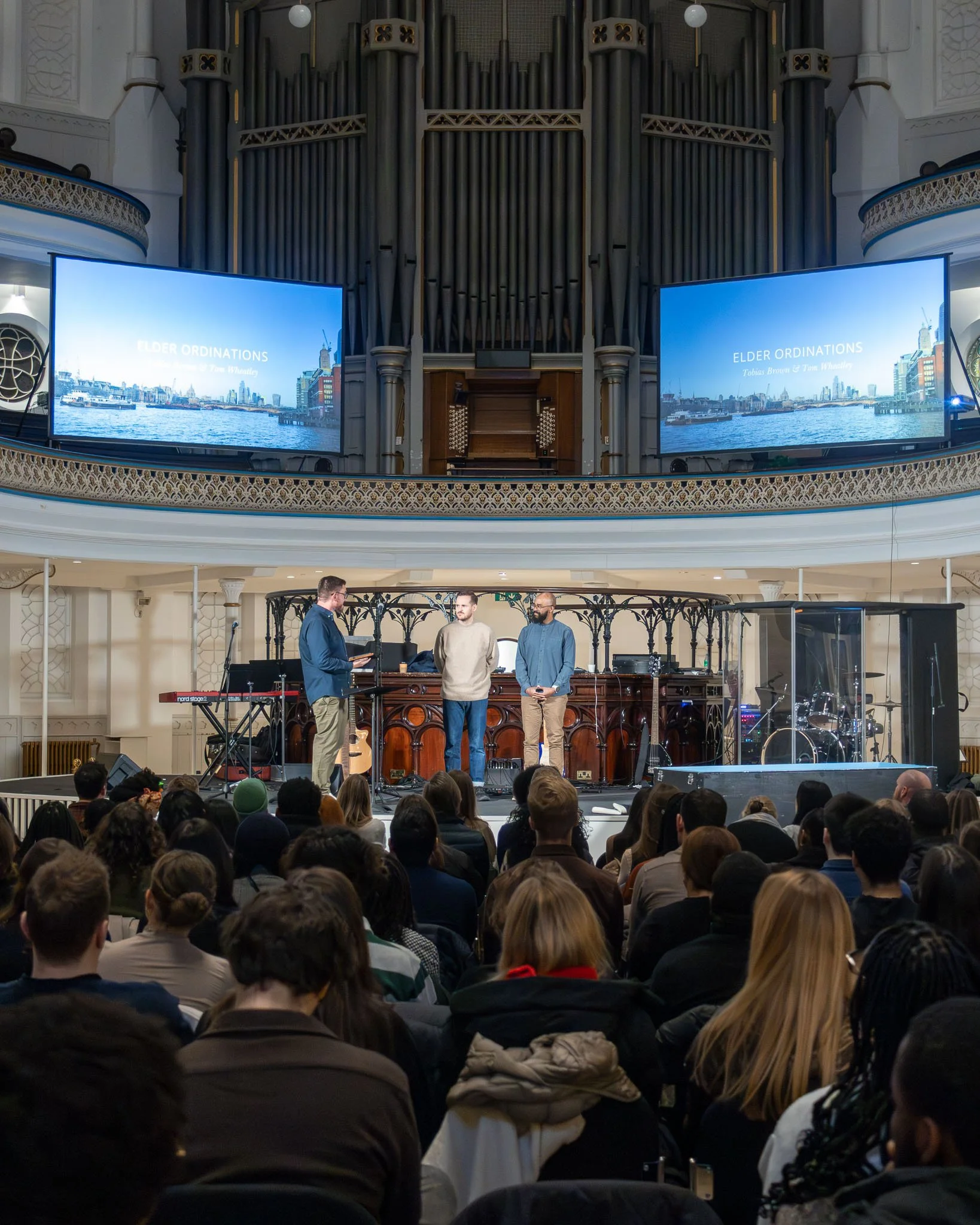 People attending a church service or event with a large congregation, three men are speaking on stage at the front, with two large screens displaying a presentation titled 'Elder Ordinations' above them. The venue features ornate architecture and a p