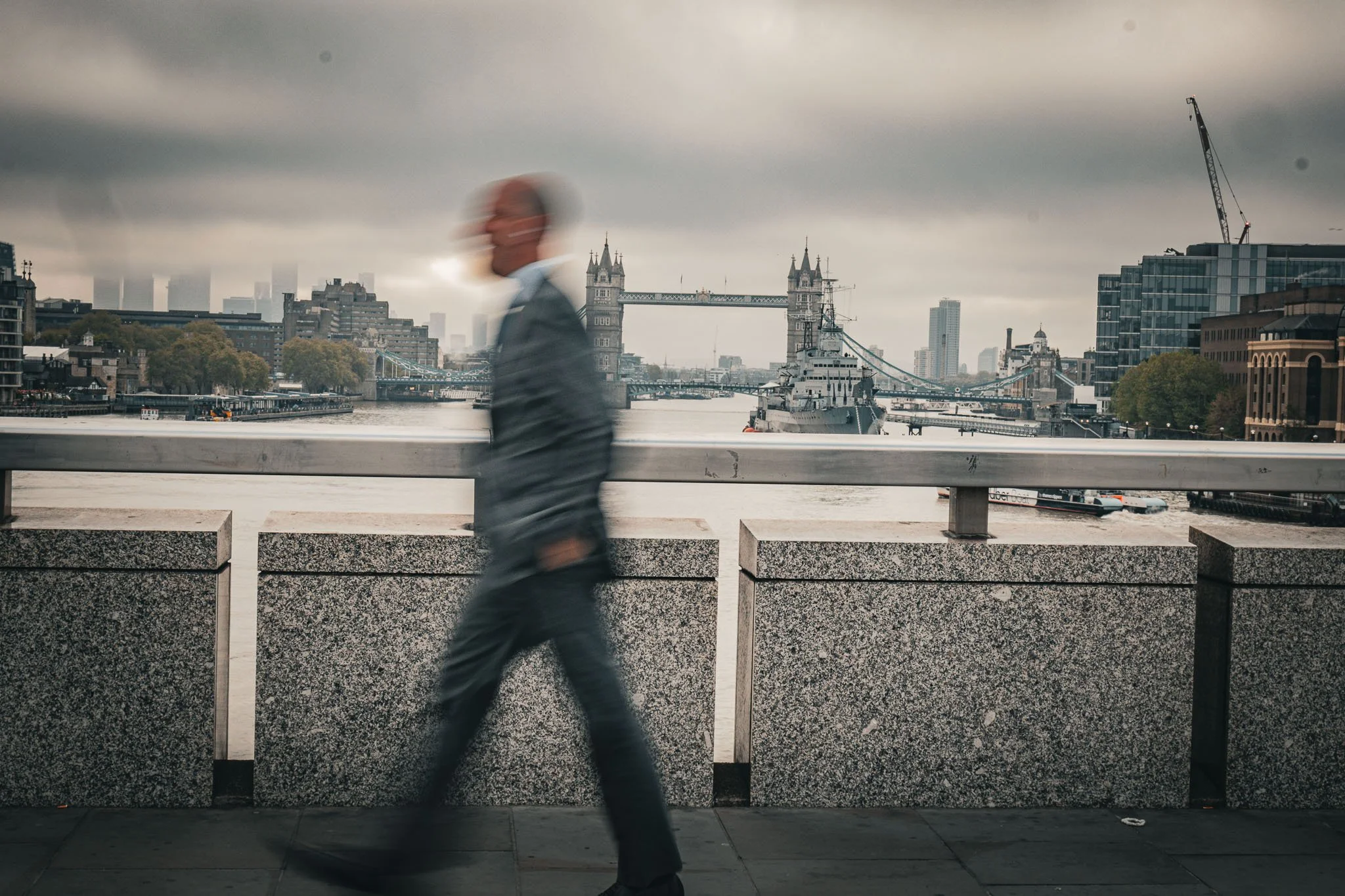 A person walking along a waterfront with the Tower Bridge and city skyline in the background on a cloudy day.