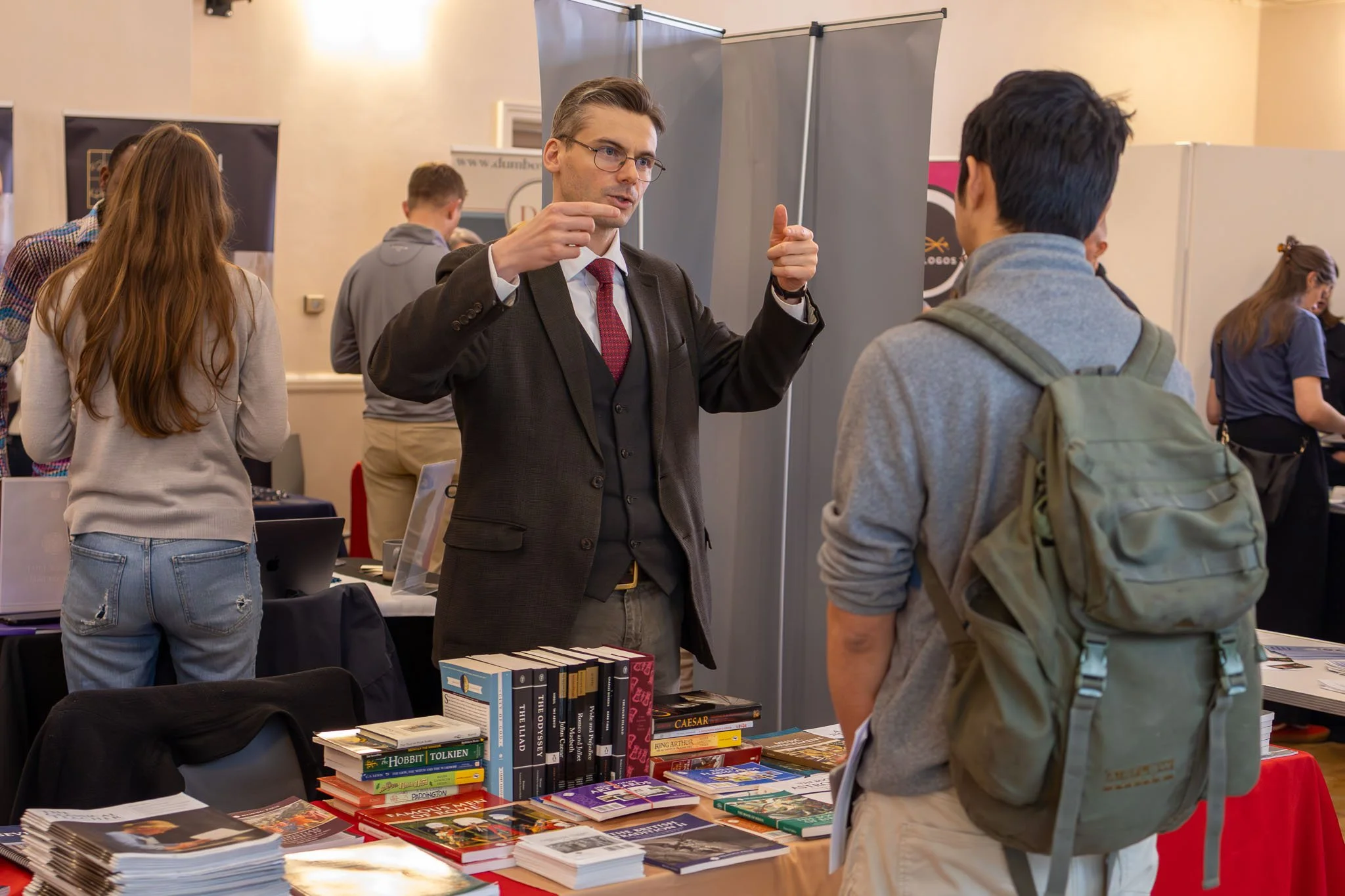 A man in a dark suit with glasses and a red tie is speaking to a young man with dark hair, wearing a gray hoodie and a backpack, at a book fair or convention. The background features other people browsing and tables filled with books and printed mate
