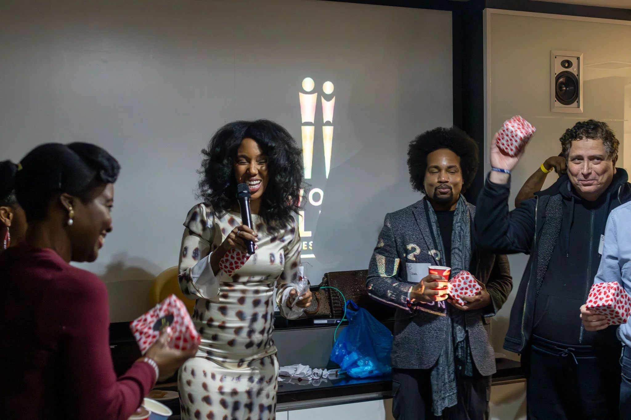 People at an event, some wearing red and white polka-dotted hats, with one woman holding a microphone and smiling, while others hold cups and interact.