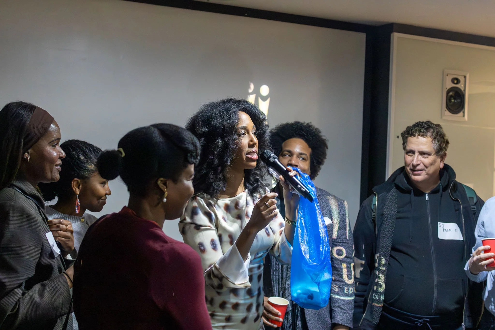 A diverse group of people gathered indoors, with one woman speaking into a microphone. The group includes women and men of different ages and ethnicities. Some are smiling, and one person holds a red cup. The setting is a singles event.