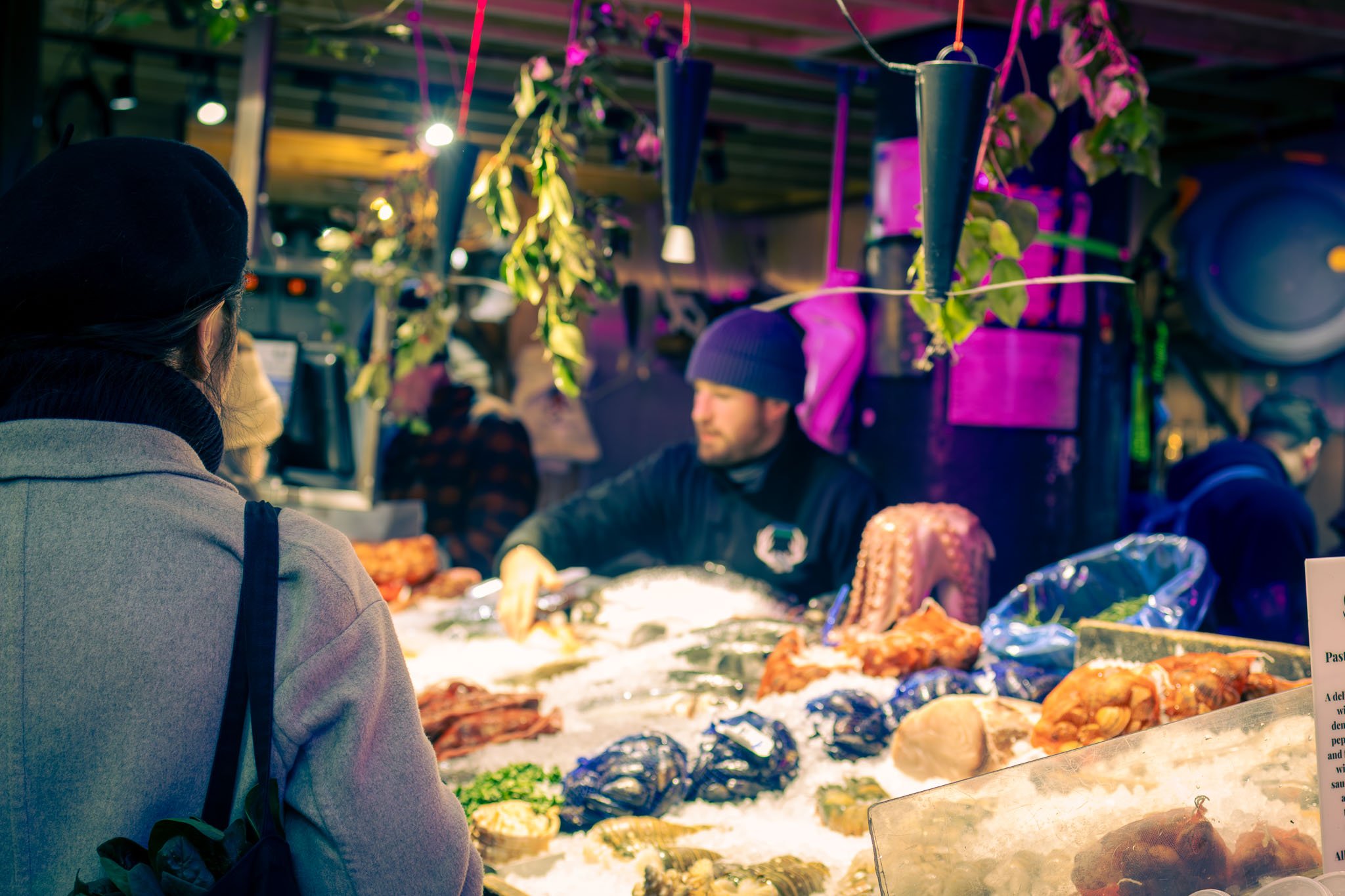 People shopping at Borough Market with a display of various cooked foods, vegetables, and hanging cone-shaped paper decorations, under colorful lighting.