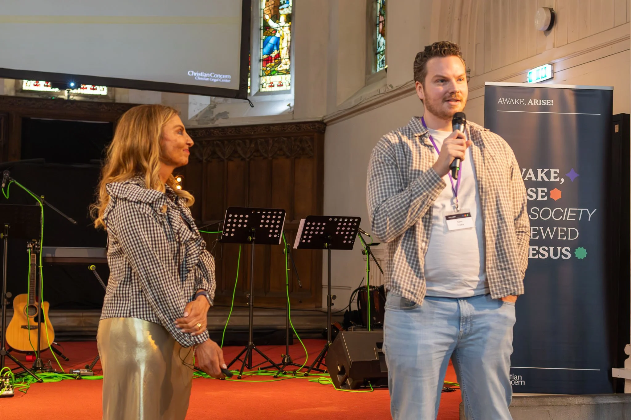 A woman and a man are speaking at a Christian event inside a church with stained glass windows. The woman is looking at the man, who is holding a microphone, while the woman has her hands clasped. There are music stands and guitars nearby, and a bann