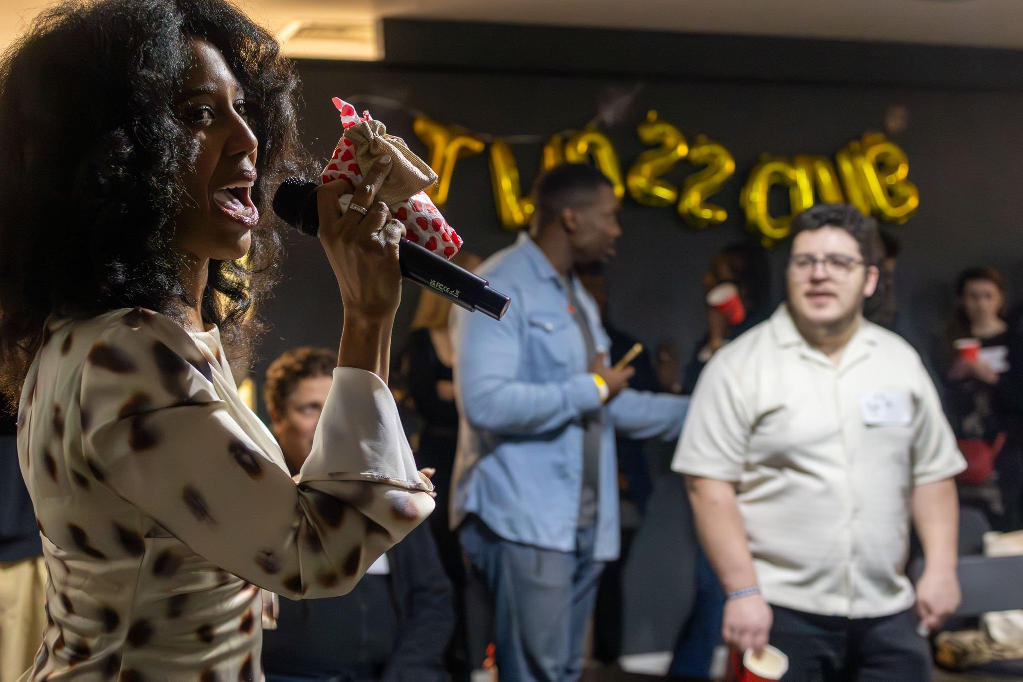 A woman with curly dark hair and a tattoo on her arm speaking into a microphone at an event with golden balloons spelling 'Two 22 Club' in the background and several people standing nearby.