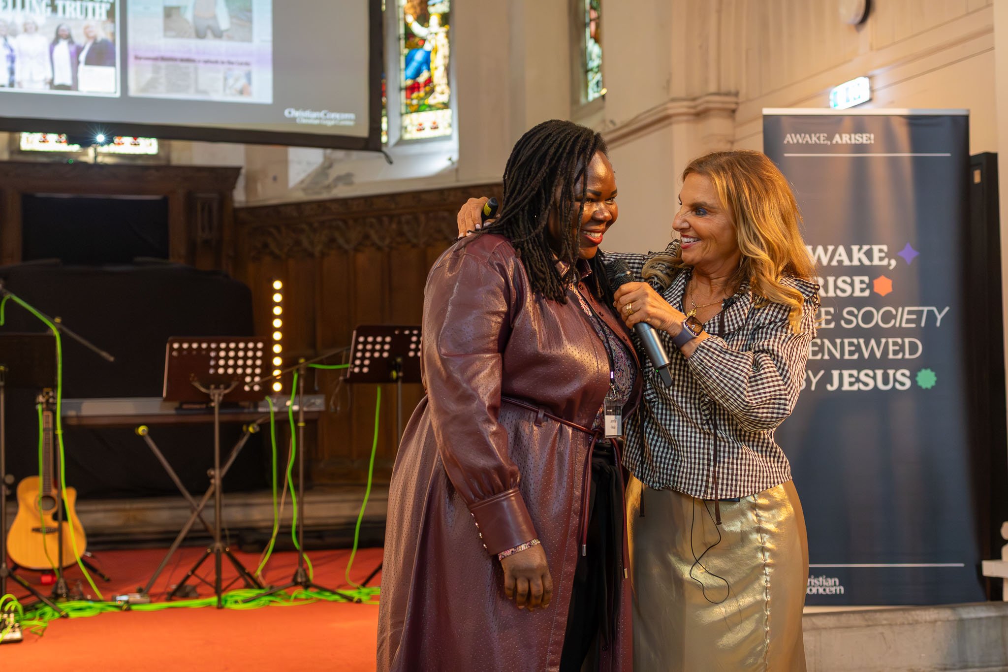 Two women are standing close together inside a church, smiling and talking, with one woman holding a microphone. One woman has long, styled red hair and is wearing a plaid shirt, while the other woman has long dreadlocks and is wearing a shiny leathe
