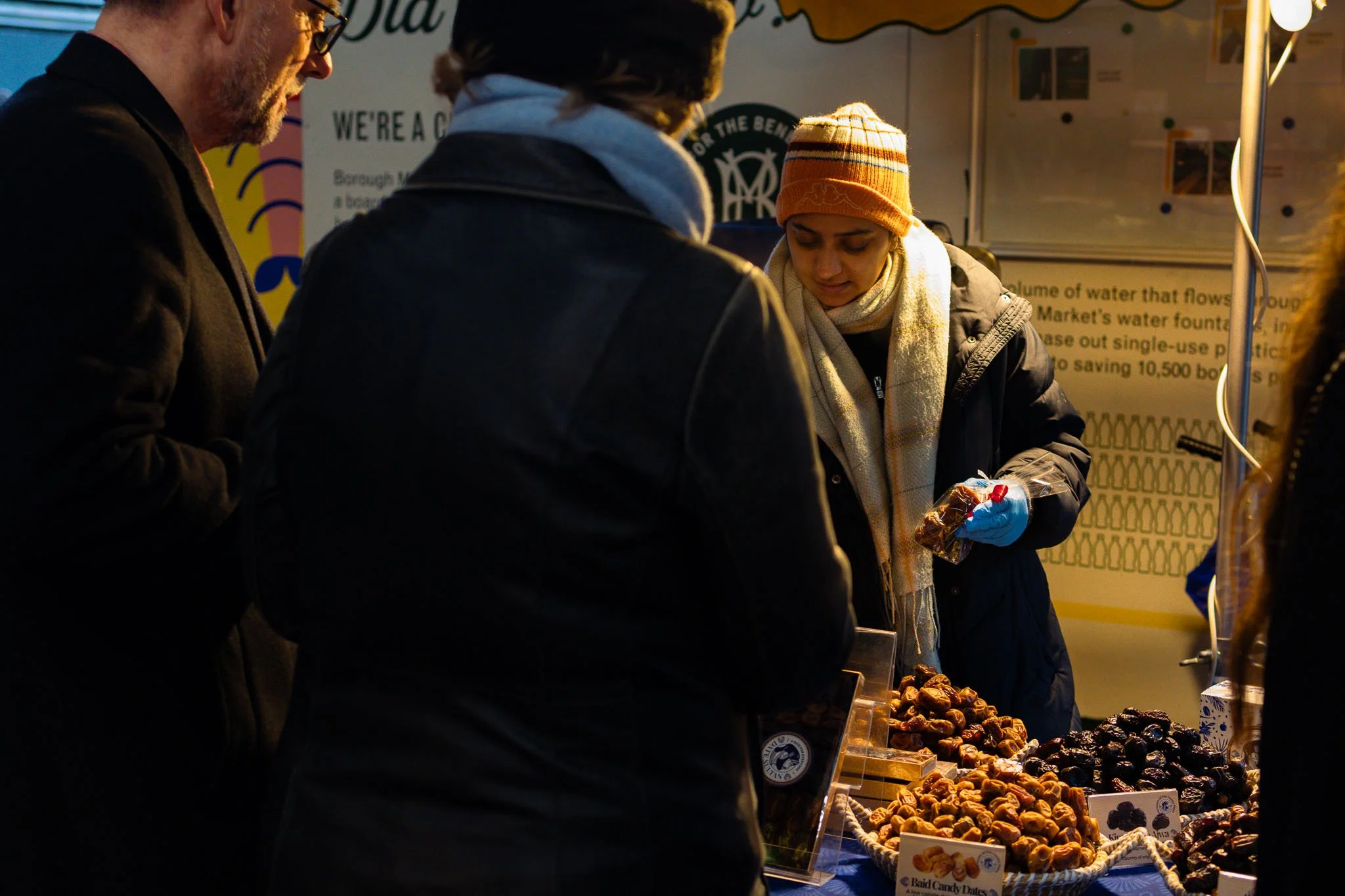 People shopping for fresh dates at Borough Market stand during morning hours, with a woman wearing a beanie and scarf, and others looking at a variety of dates displayed on the counter.