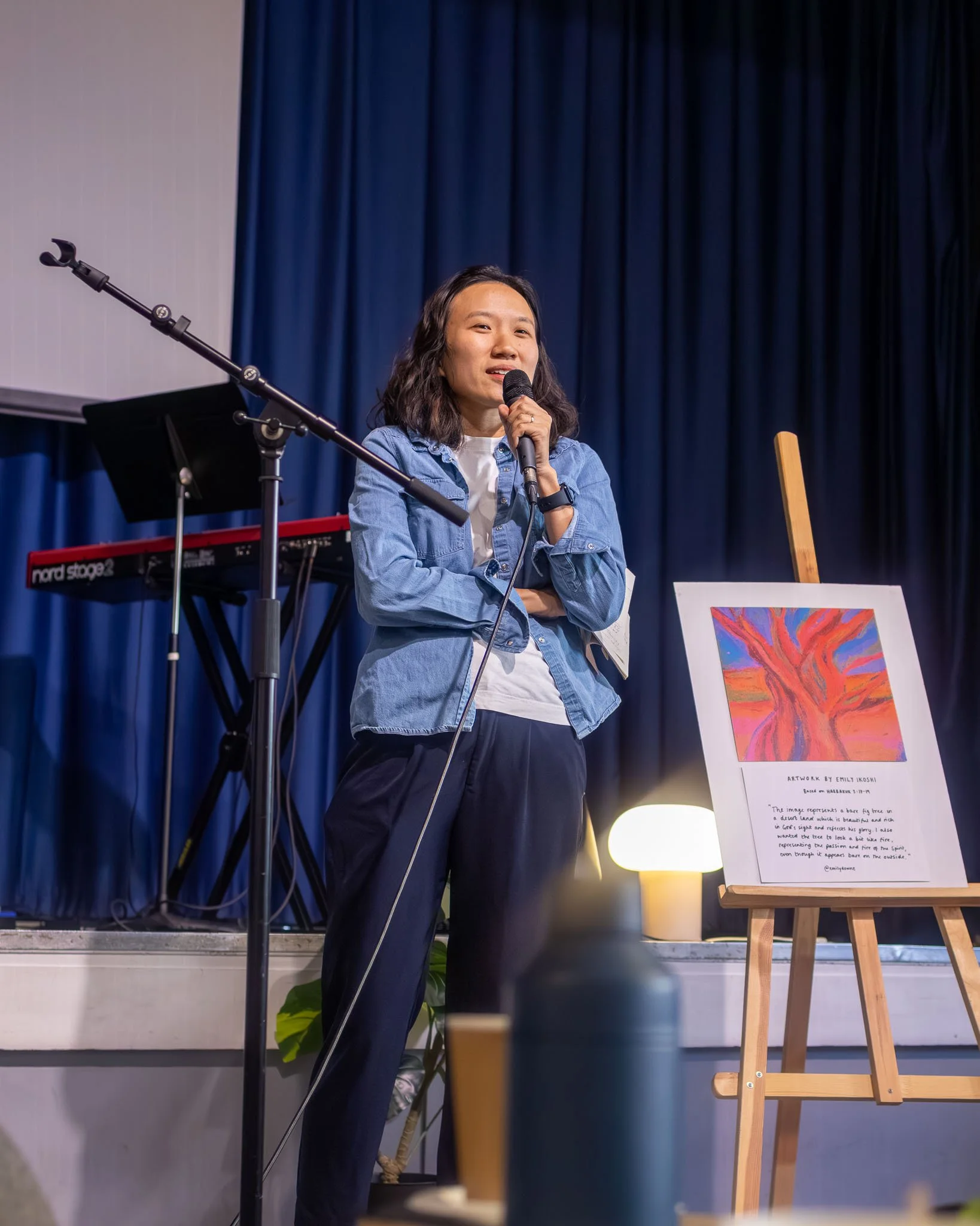 A woman with shoulder-length dark hair, wearing a denim jacket and white shirt, stands on stage holding a microphone. Behind her is a blue curtain, and to her right is an easel with colorful artwork. Part of a keyboard and a music stand are visible i