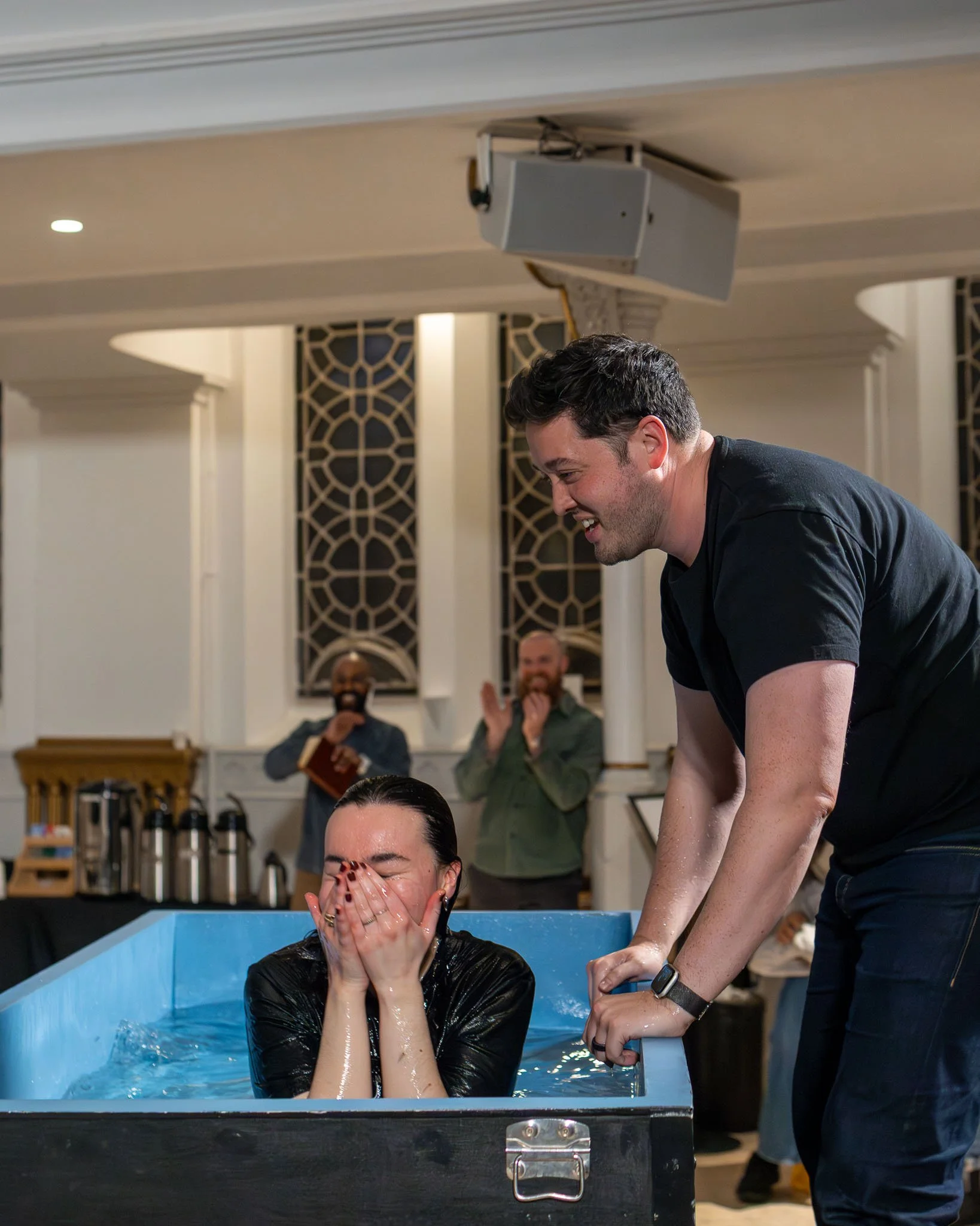 A woman is being baptized in a small indoor pool filled with water, with her hands covering her face. A man is standing beside the pool, smiling and holding her arm. In the background, two people are clapping and smiling, one holding a Bible, in a ro