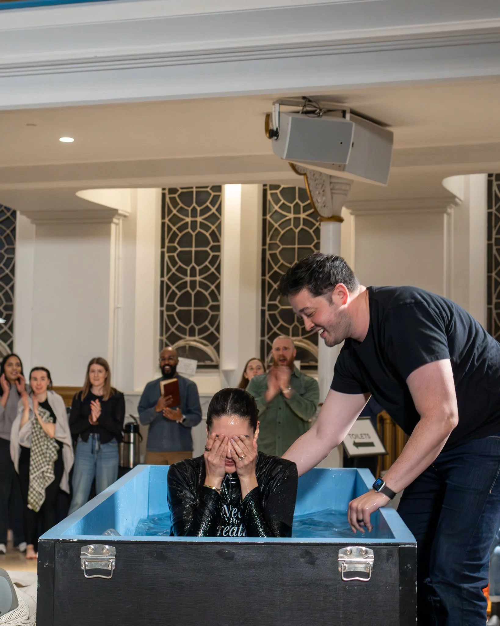 A woman is sitting in a baptismal font with water, covering her face with her hands. A man is standing next to her, holding her arm and smiling. Several onlookers are standing behind them, watching and reacting with smiles and applause in a bright ro