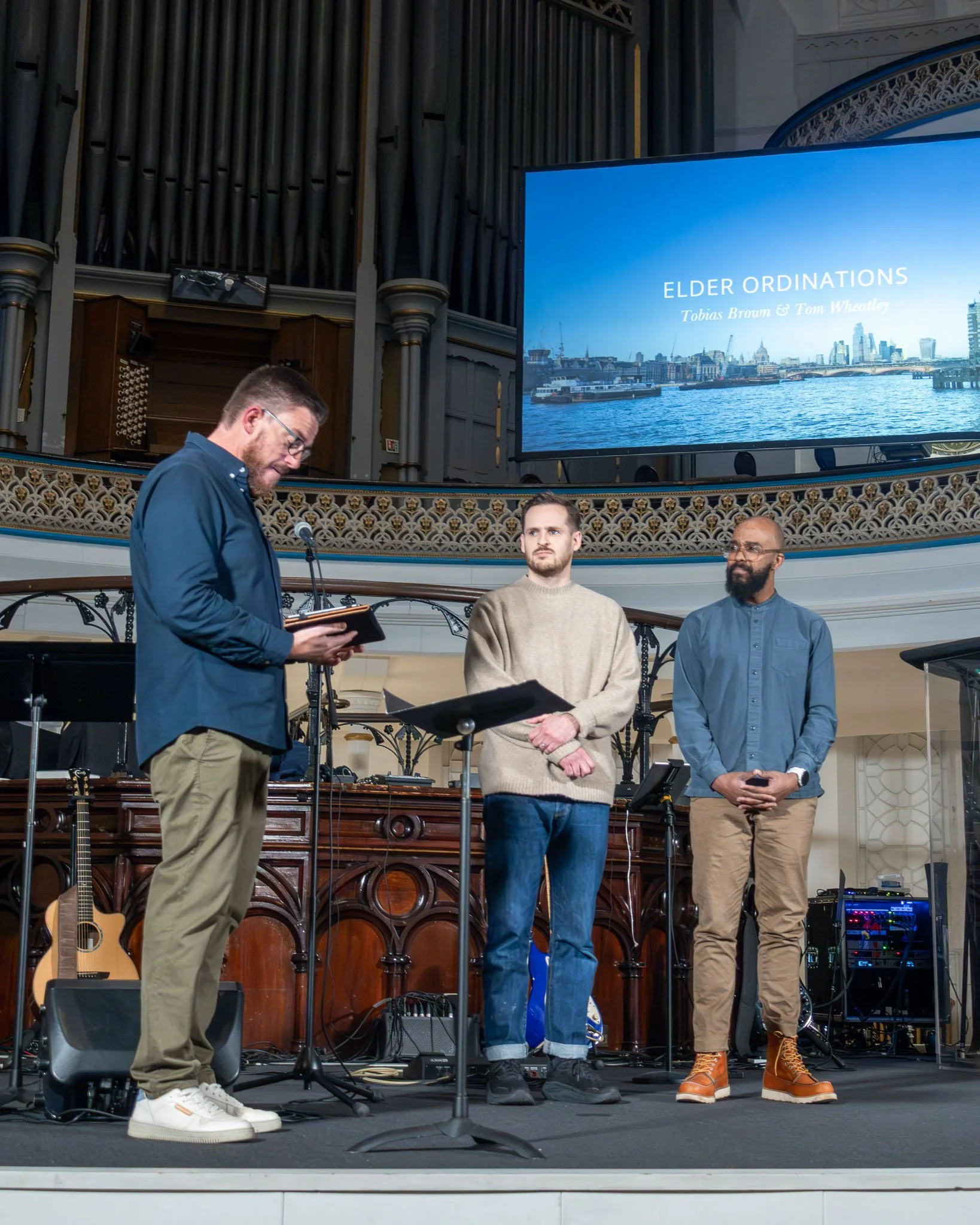 Three men standing on a stage in a church or chapel, with a large pipe organ in the background. One man is reading from a book, the other two are listening. A large screen displays "ELDER ORDINATIONS, Tobias Brown & Tom Wheatly" with a cityscape imag