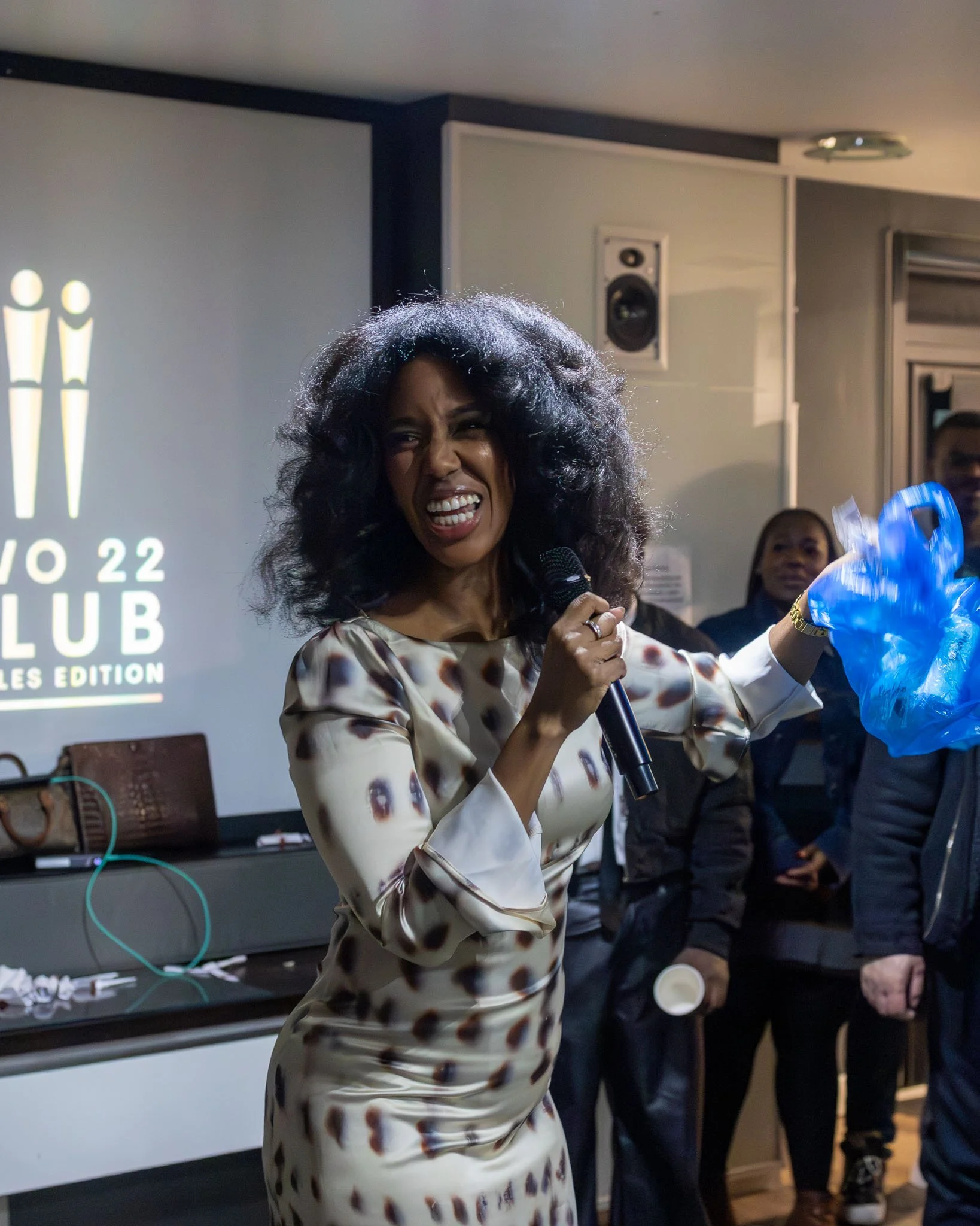 A woman with curly black hair wearing a white and brown animal print dress speaking into a microphone at an indoor event with a black speaker and audience members in the background.
