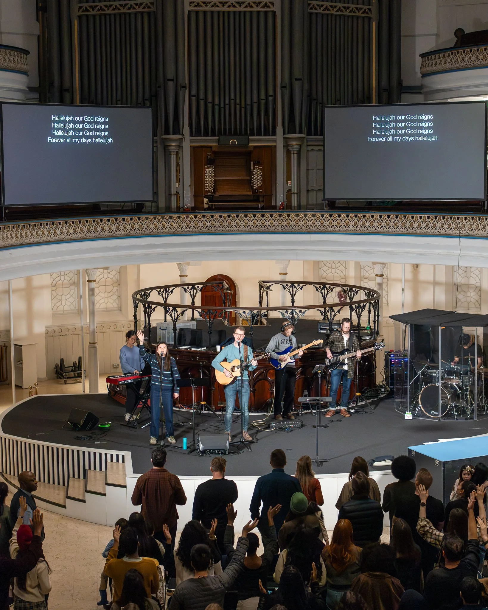 A church with a band performing. The band includes a woman singing at a microphone, a man playing acoustic guitar, another man playing electric guitar, and a woman at a keyboard. Audience members are standing, raising their hands, and participating i