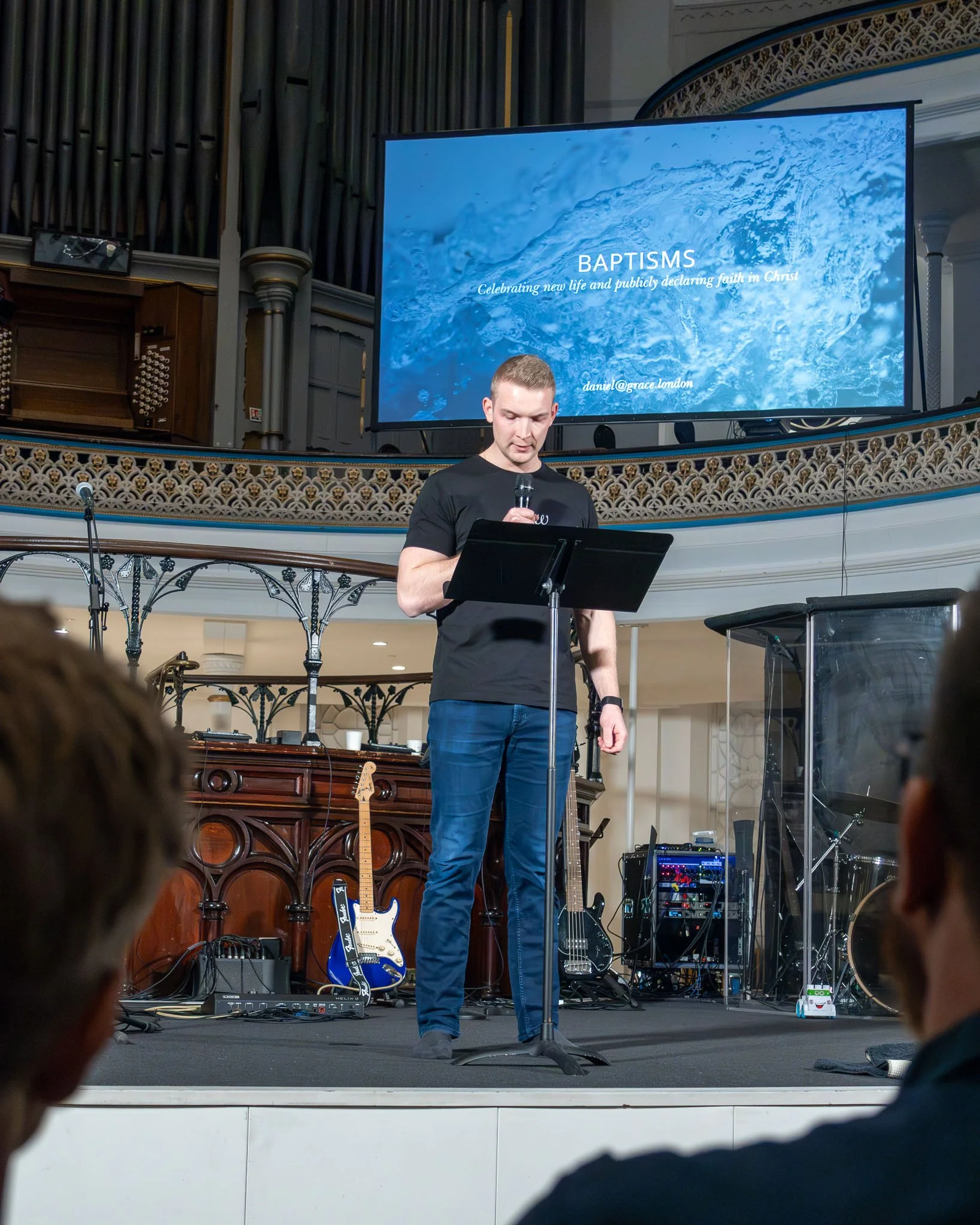 A man standing on stage holding a microphone, with musical instruments and equipment behind him. A large screen displays the text 'BAPTISMS' and a background of water, with additional smaller text related to a church event.