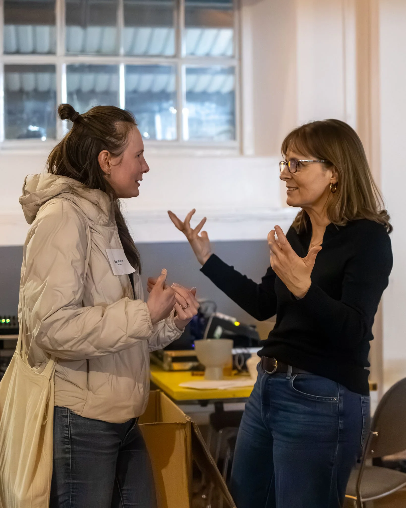 Two women are engaged in a conversation indoors, with one woman wearing glasses and a black top, and the other in a beige jacket. They appear to be talking animatedly.