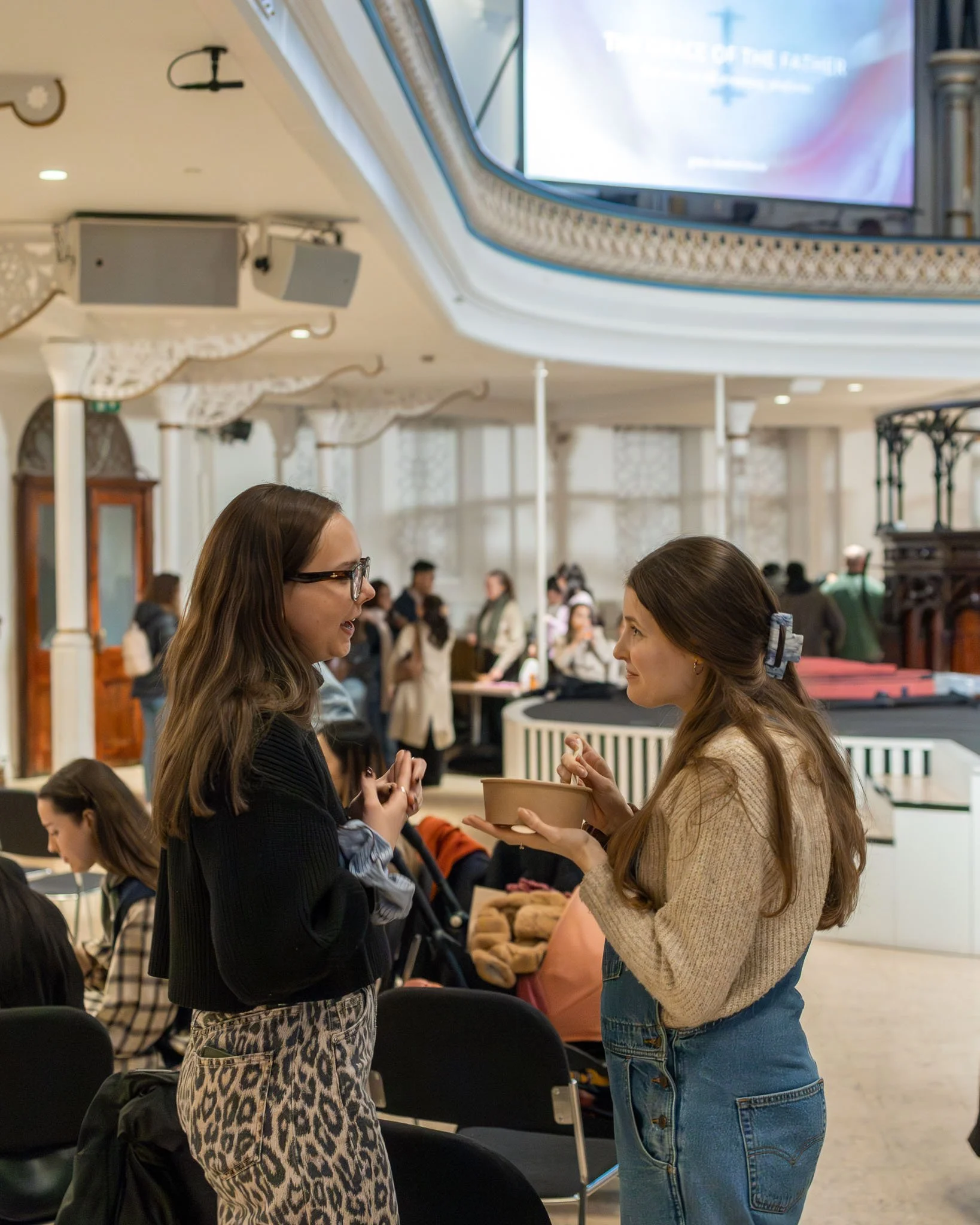 Two women are engaged in conversation in an indoor setting, with one woman offering a bowl to the other. The background shows other people and a large screen on the wall.