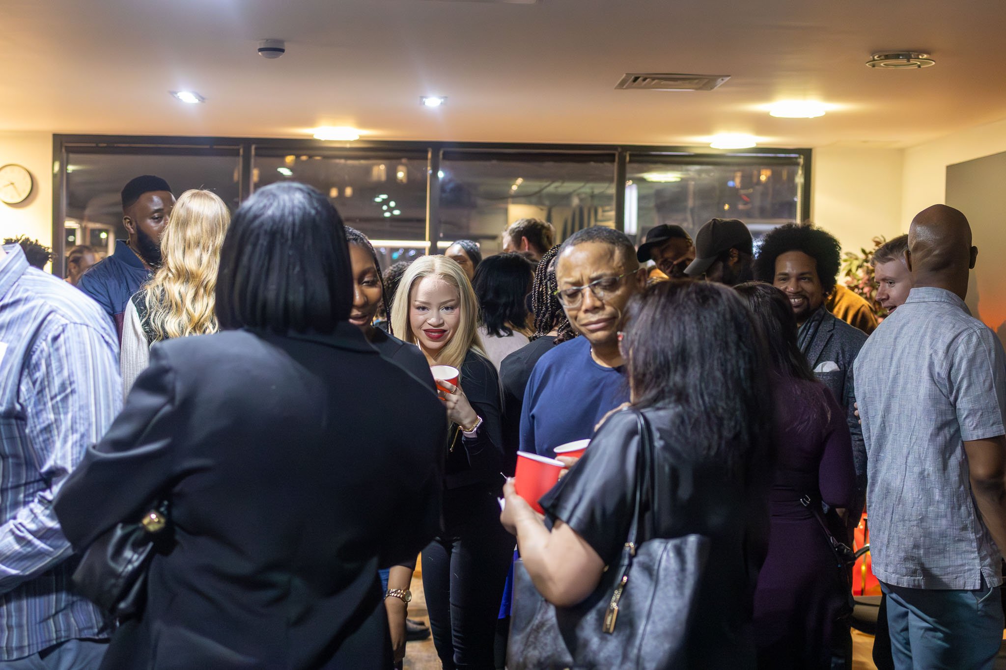 Group of diverse people at an event, holding red cups, engaging in conversations indoors with warm lighting.