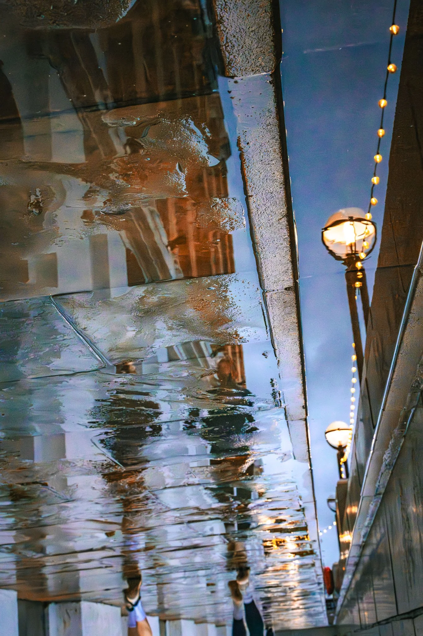 Indoor hallway with wet, reflective floor and a row of decorative string lights hanging along one side. A person with white shoes can be seen walking on the floor.