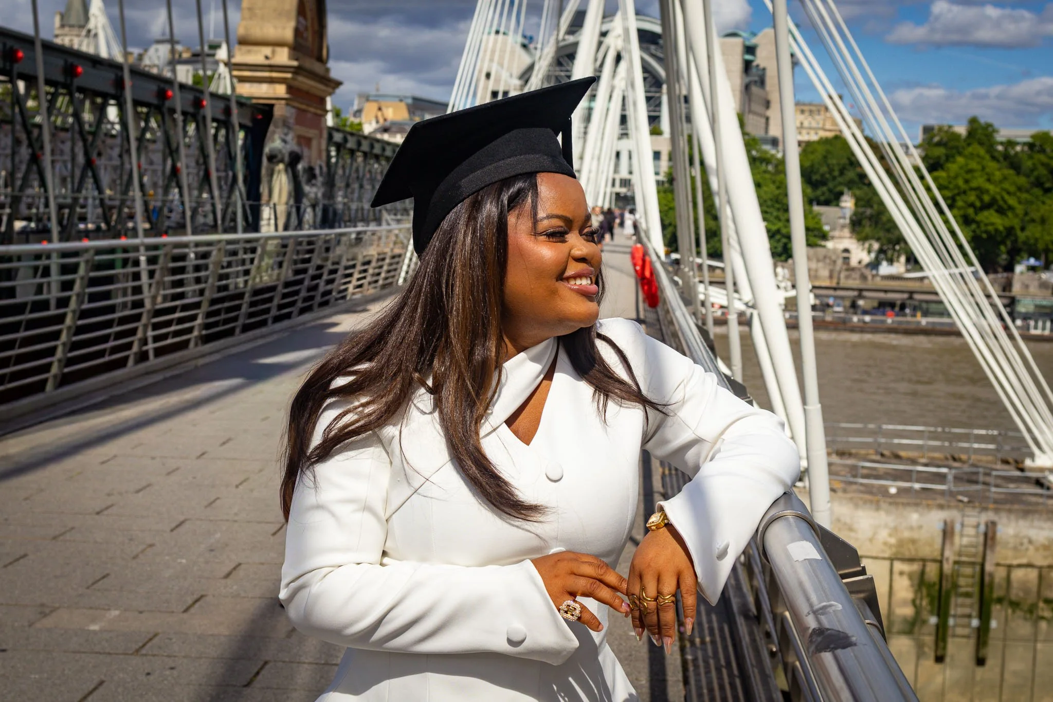 Woman in white blazer and black graduation cap leaning on bridge railing outdoors on a sunny day.