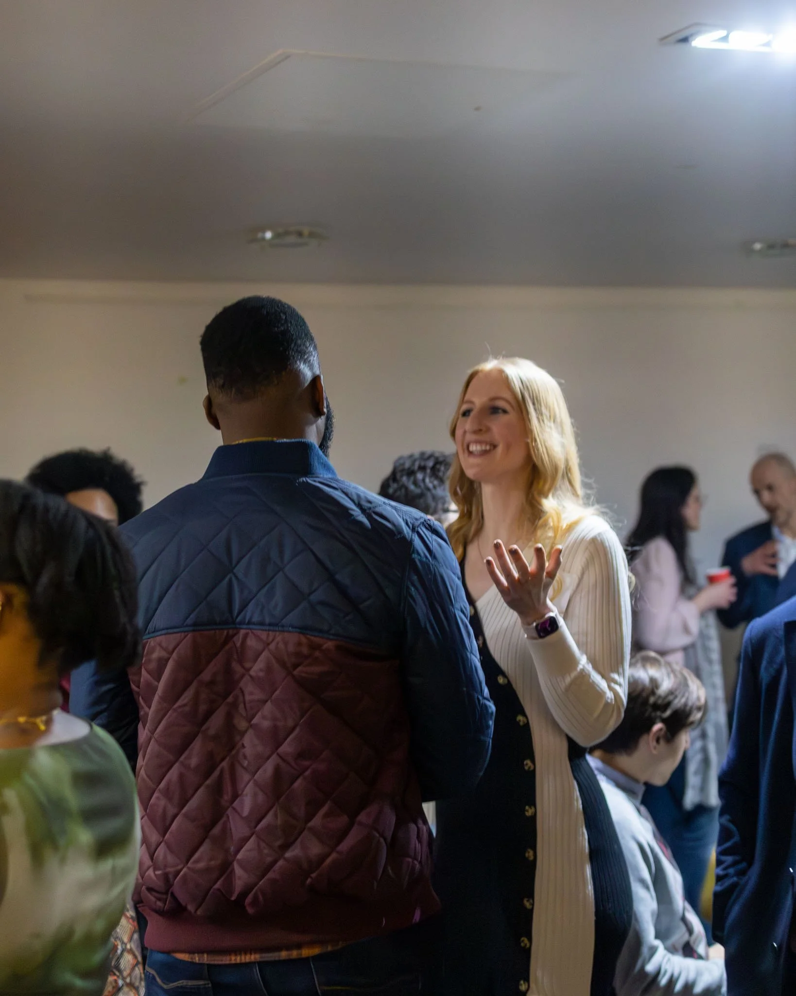 People gathered in a gathering or social event, with some engaged in conversation. The woman in the foreground is smiling and gesturing with her hand, and others are seen in the background talking. The ceiling has a vent or smoke detector, and the ro