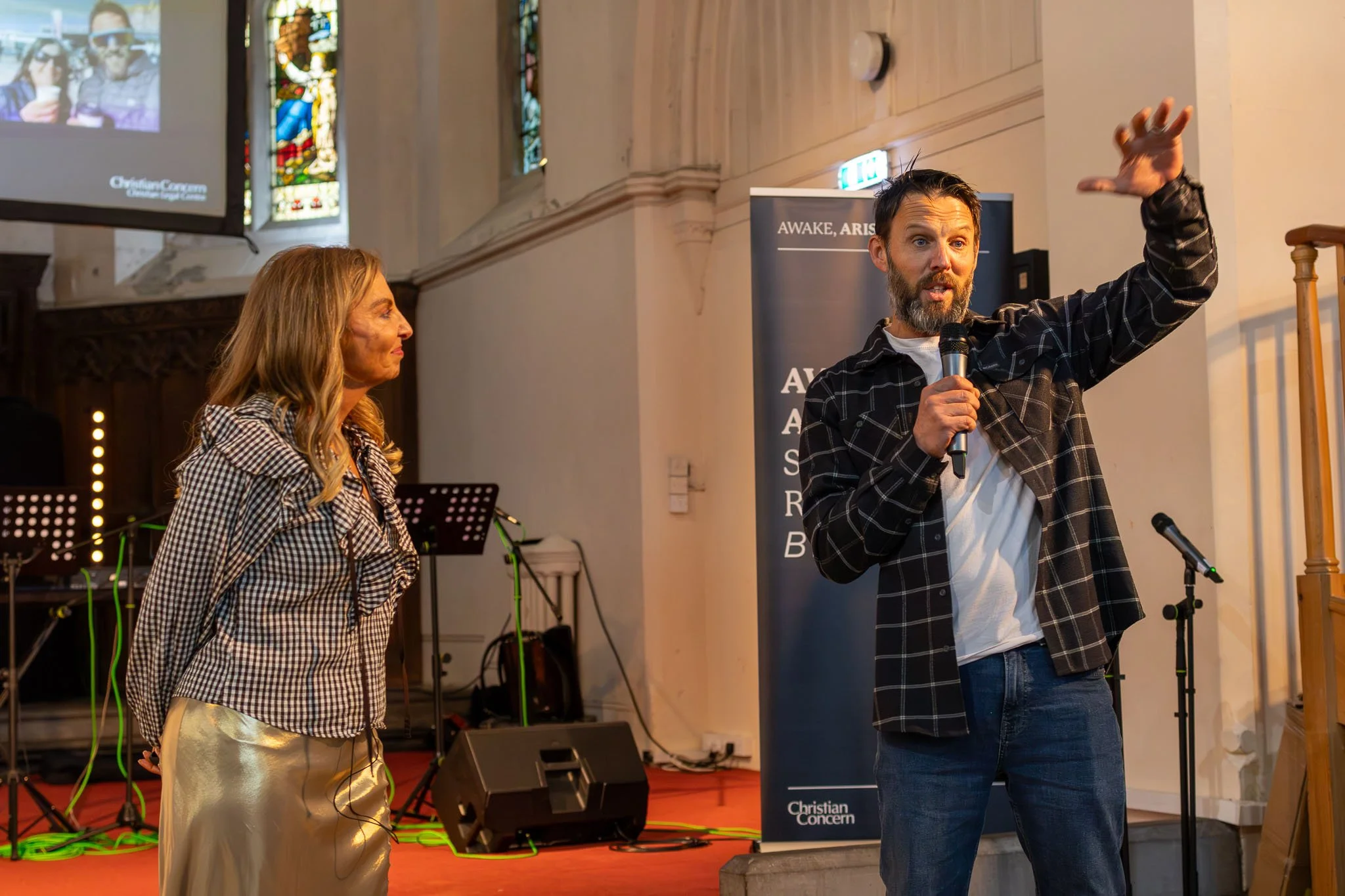 A man with a beard and short hair, dressed in a dark plaid shirt and white T-shirt, is speaking into a microphone and raising his left hand. A woman with long wavy hair, wearing a checkered blouse and shiny gold pants, is standing next to him, listen