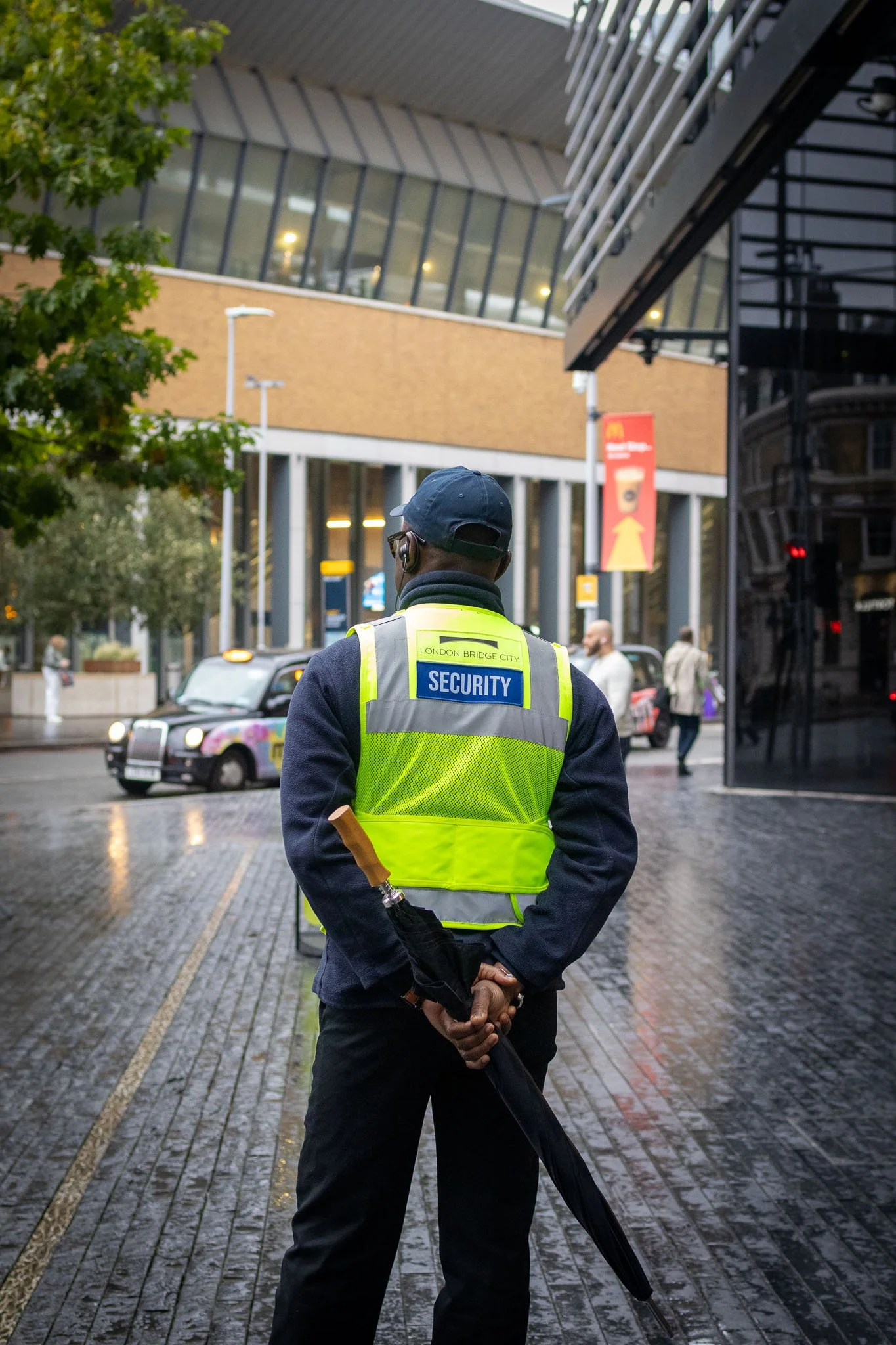 A security guard wearing a yellow vest that says 'SECURITY' on the back, standing outdoors on a wet brick sidewalk near London Bridge, with people walking nearby and a vintage taxi cab in the background.