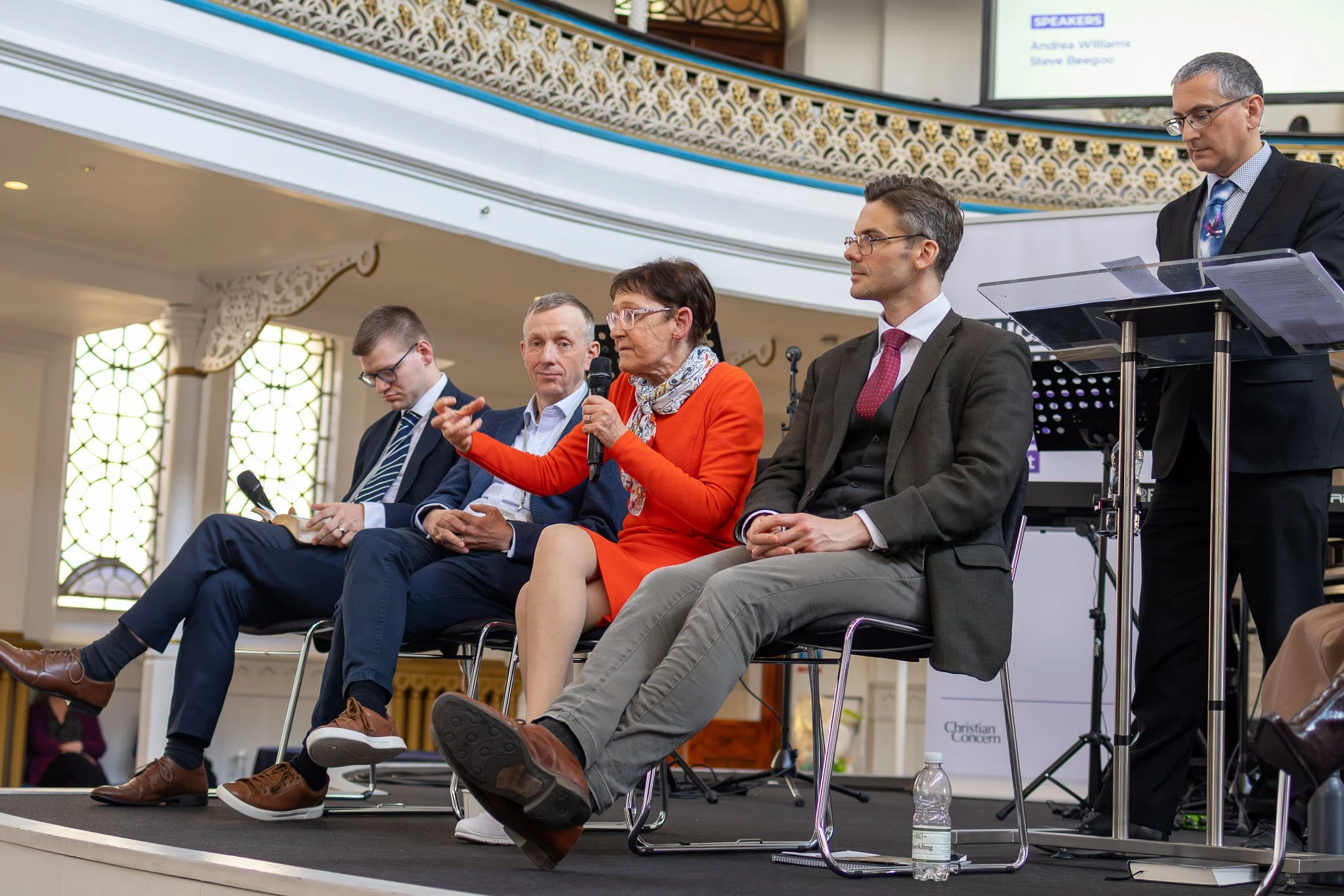 A panel discussion taking place in a large, ornate room with stained glass windows. Five people are seated on stage, four men and one woman, who is speaking into a microphone. Behind them is a screen displaying text, and there is a clear podium with 