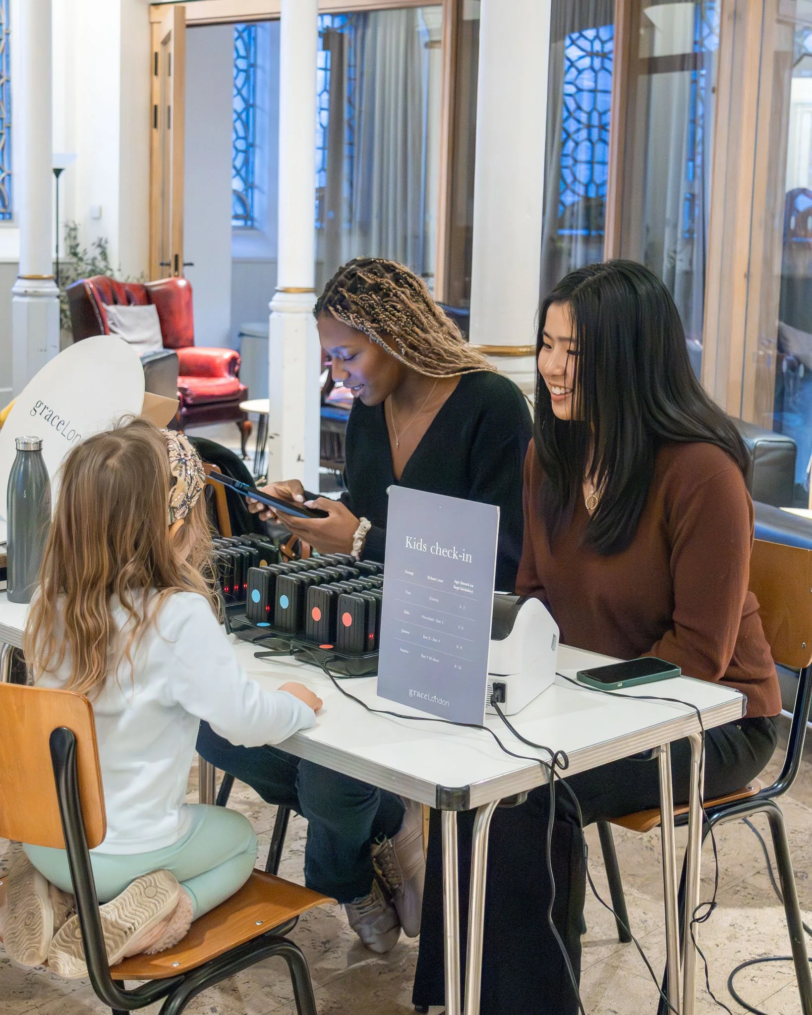 People at a kids' check-in counter with electronic devices in a well-lit indoor setting, with wooden chairs, glass windows, and decorative wall panels in the background.