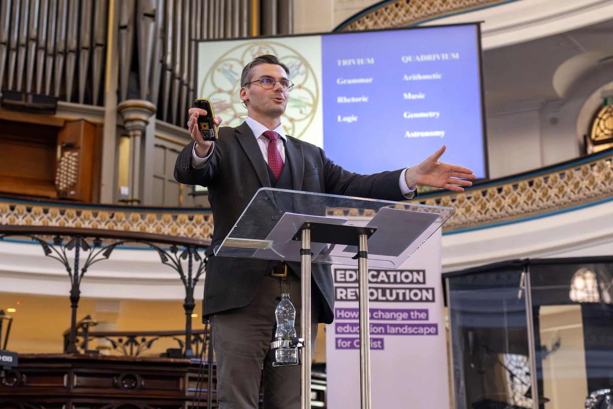 A man in a suit giving a presentation at a podium with a large screen behind him displaying a slide about education reform.