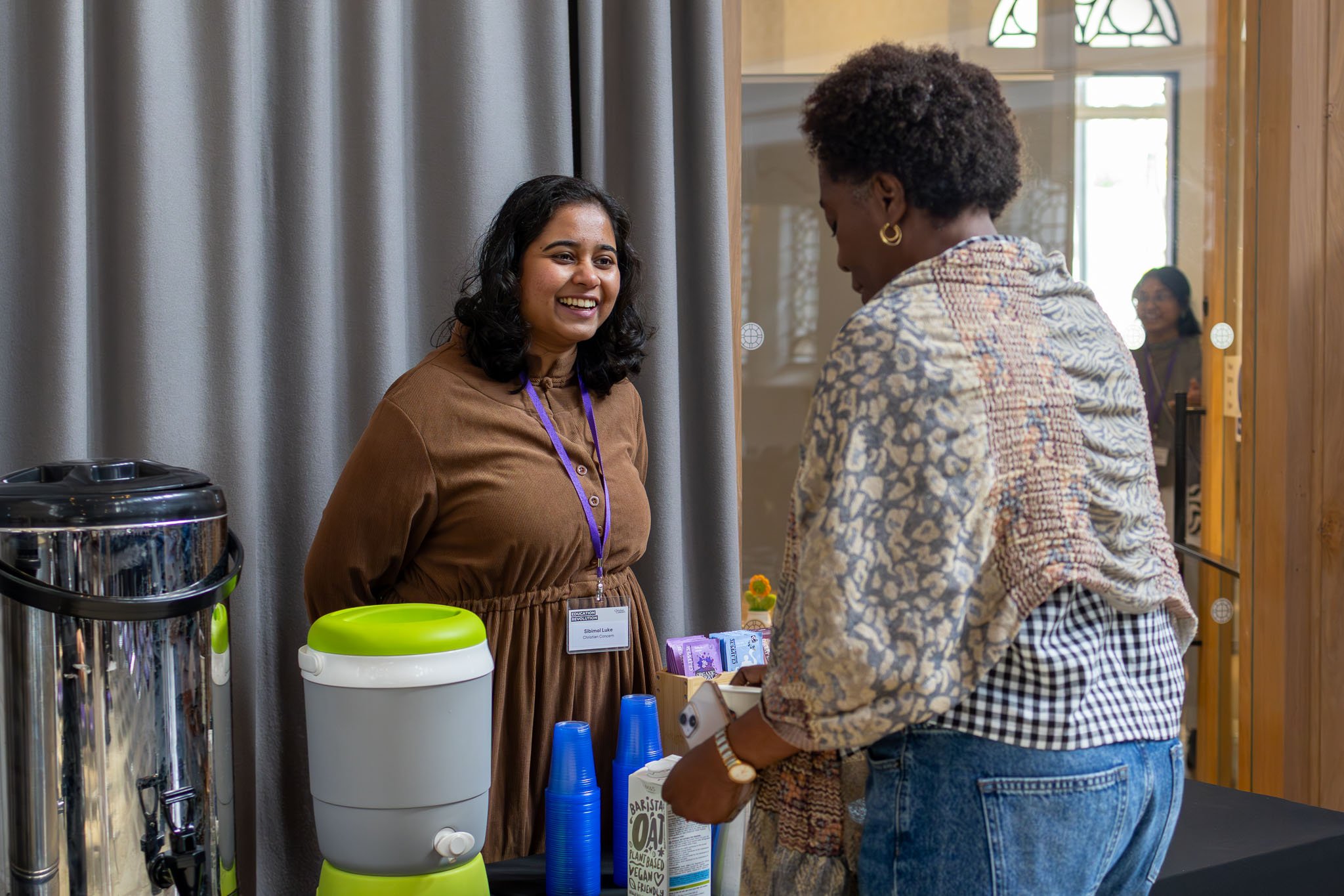 Two women having a conversation at a booth during an event. The woman on the left is smiling and wearing a brown dress with a purple lanyard. The woman on the right has curly hair, is wearing a patterned jacket and jeans, and is facing the woman on t