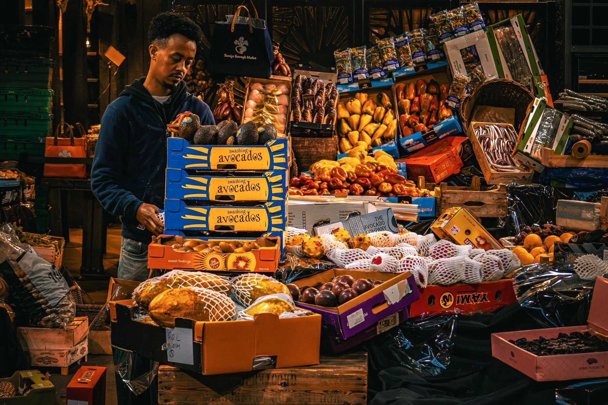 A man setting up food at Borough Market with various fruits and vegetables, including avocados, peppers, and melons, displayed on wooden tables and in boxes.