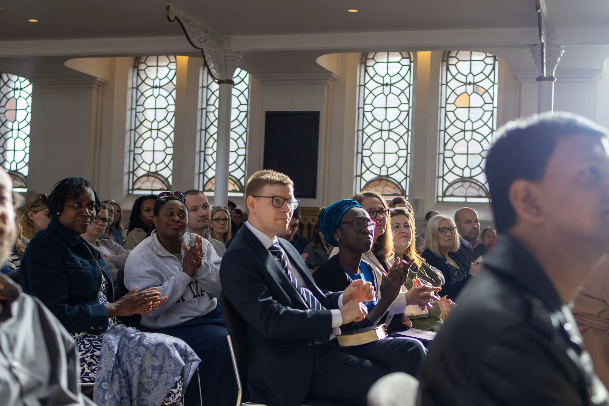 A diverse group of people sitting in pews attending a religious service or ceremony inside a church with large, decorative windowpanes.