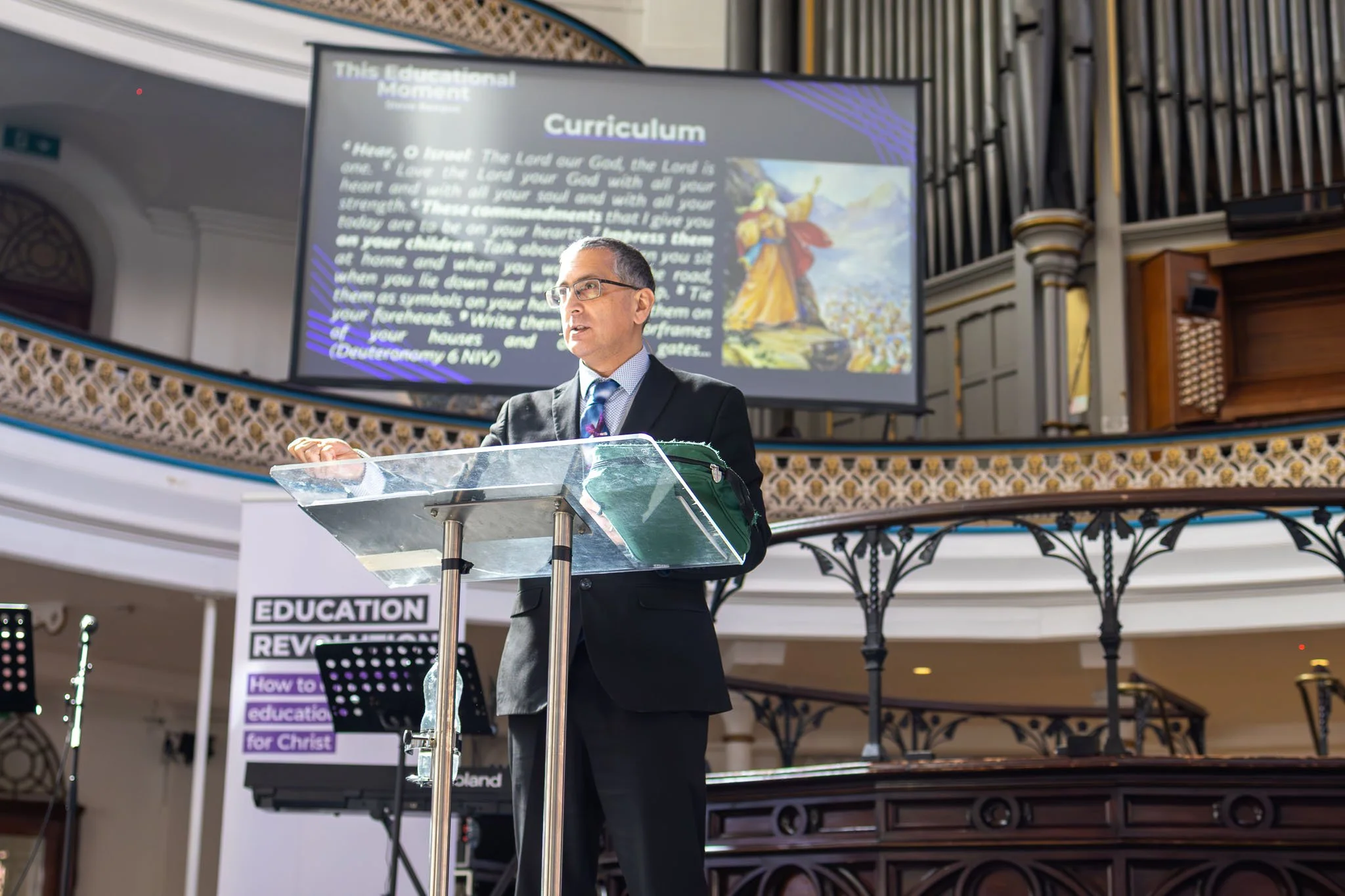 Man in a suit and glasses speaking at a podium with a sign reading 'Education' in a church or cathedral-style venue, with a large screen behind displaying text about the curriculum and an image of a biblical scene.