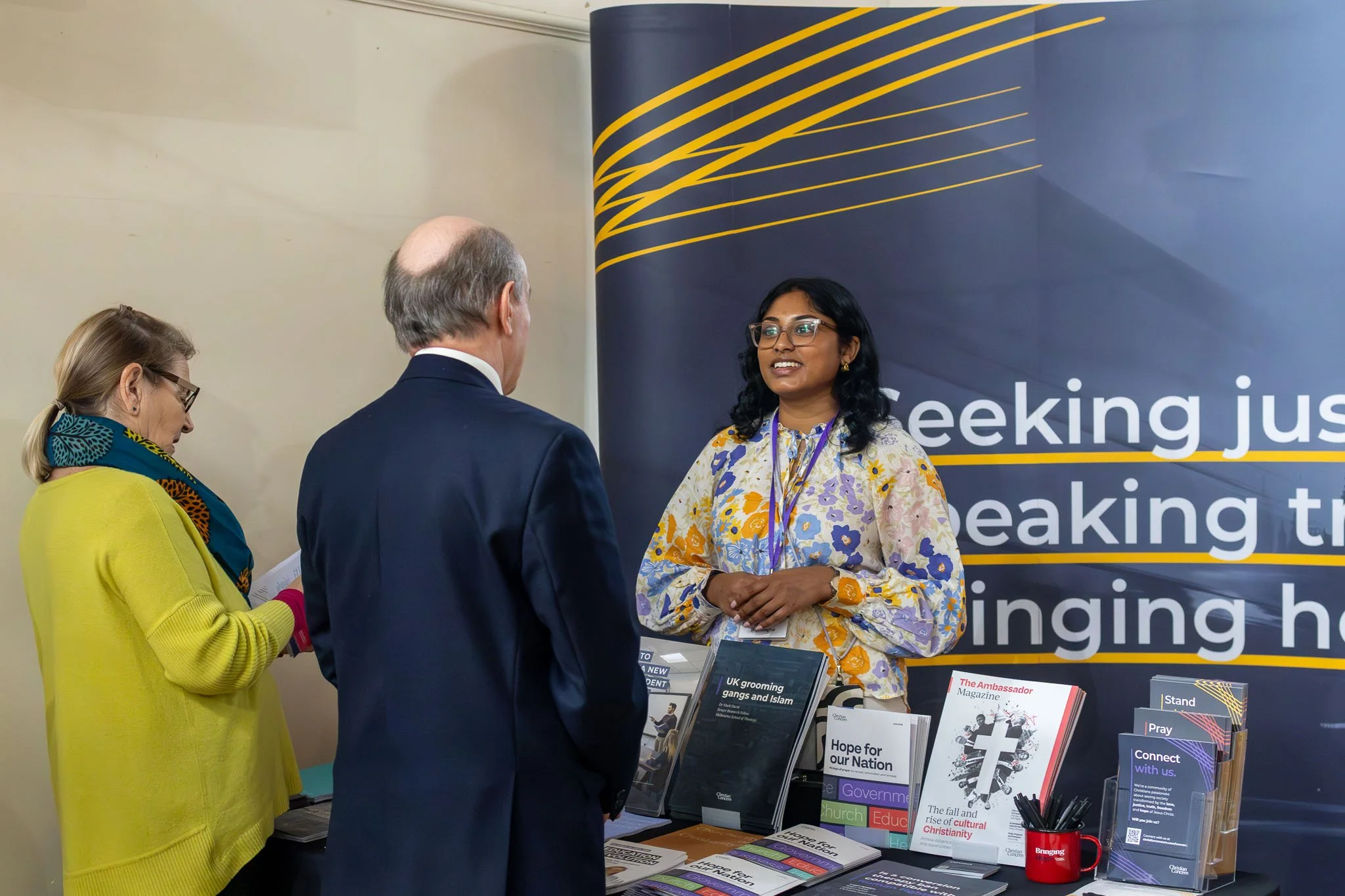 Three people standing and talking at an informational booth with brochures and a large backdrop reading 'speaking' and related words, displaying books and flyers about Christian themes.