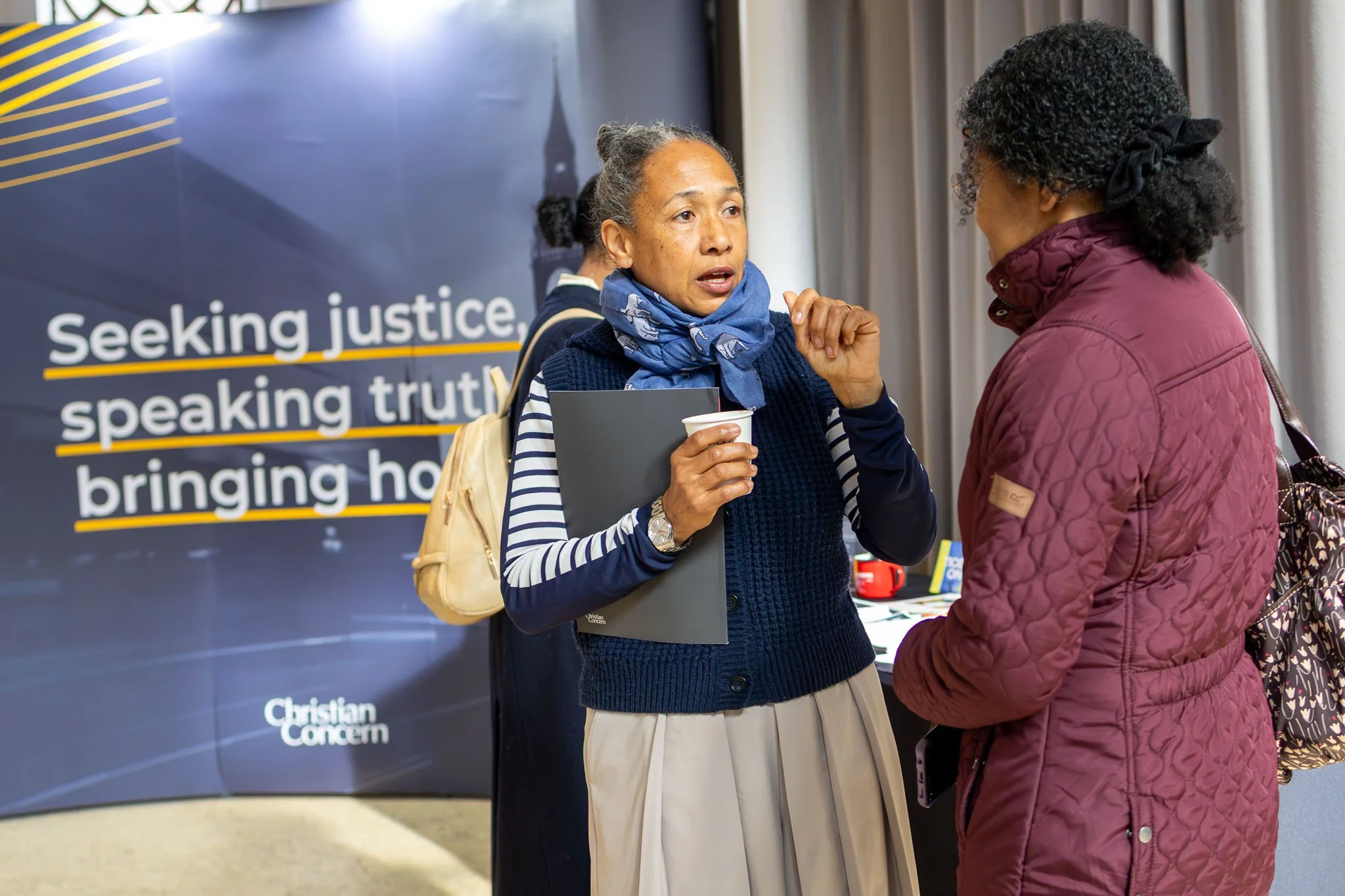 Two women are engaged in conversation at an indoor event, with a large banner in the background that reads 'Seeking justice, speaking truth, bringing ho...'. One woman has dark curly hair, a purple quilted jacket, and a bag, while the other has gray 