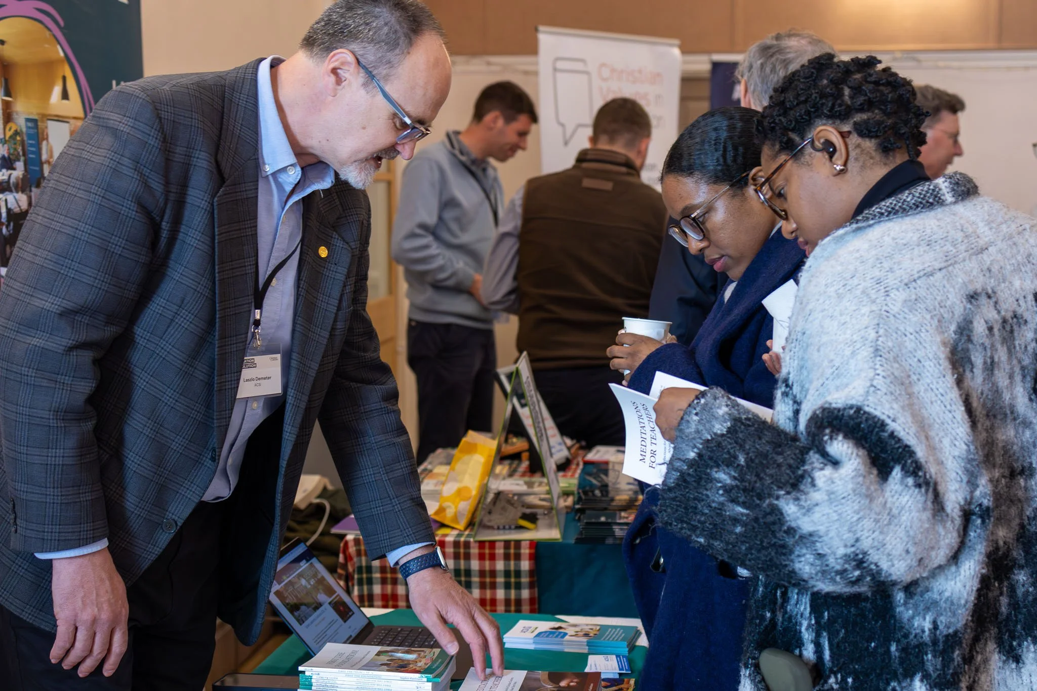 A man with glasses and a plaid suit pointing at brochures on a table while talking to two women at a conference or event. The women are holding informational pamphlets and appear engaged in conversation. Several people are visible in the background.