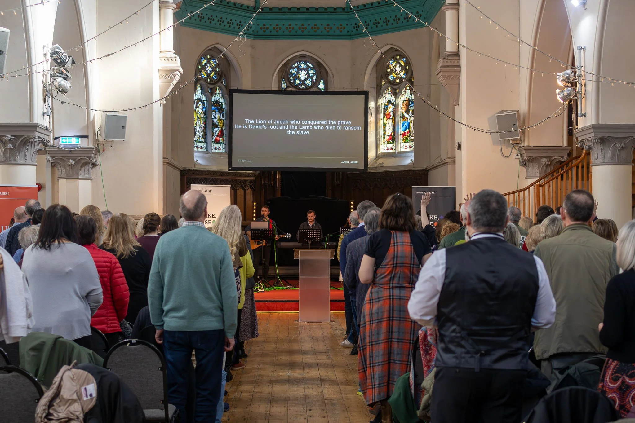 Crowd attending a church service or event, facing a stage with a large screen displaying religious text about the Lion of Judah. The church interior features stained glass windows and string lights.