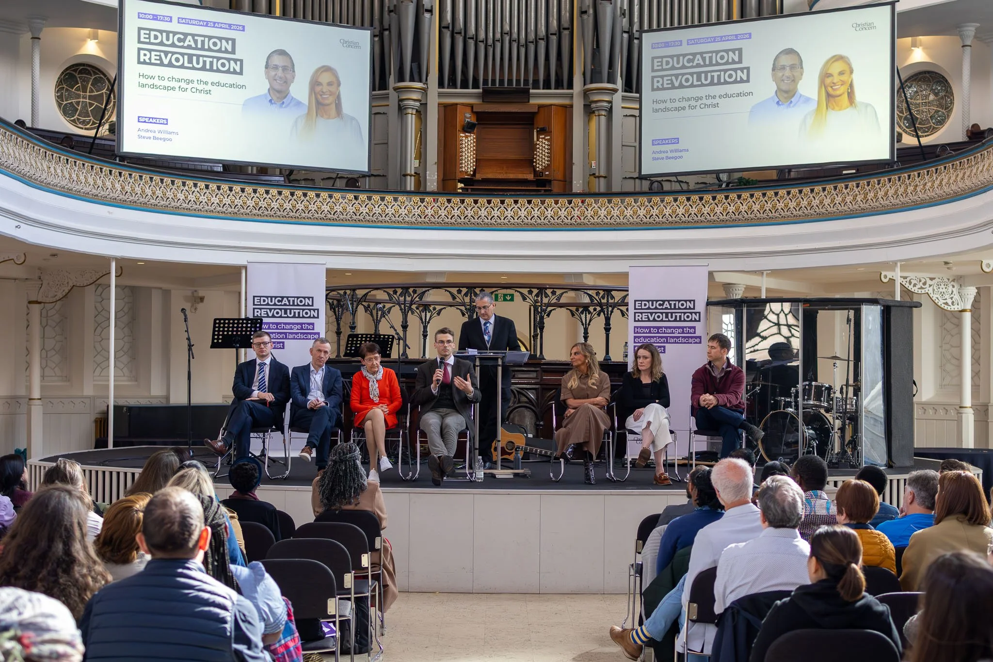 A panel discussion at a conference titled 'Education Revolution' with speakers Andrea Williams and Steve Beegoo. The event is held in a spacious hall with an audience seated in front and a large screen displaying the event details. The panel includes