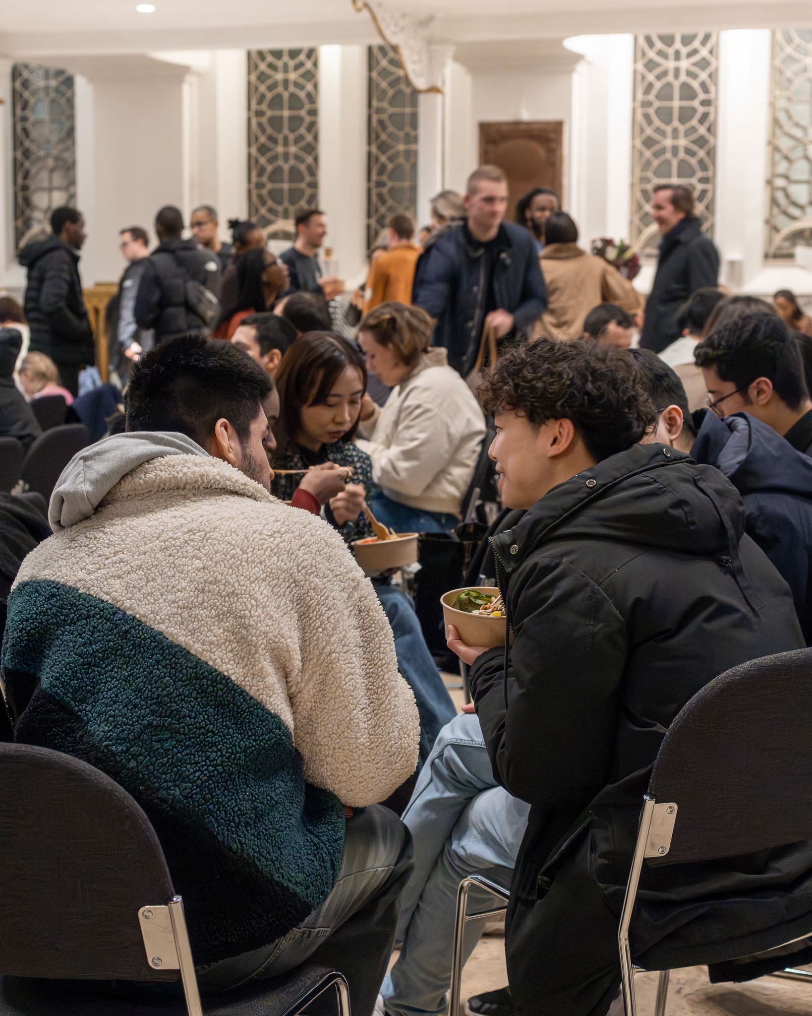 People sitting and standing in a crowded indoor event space, some eating from bowls, with decorative wall panels and a wooden door in the background.