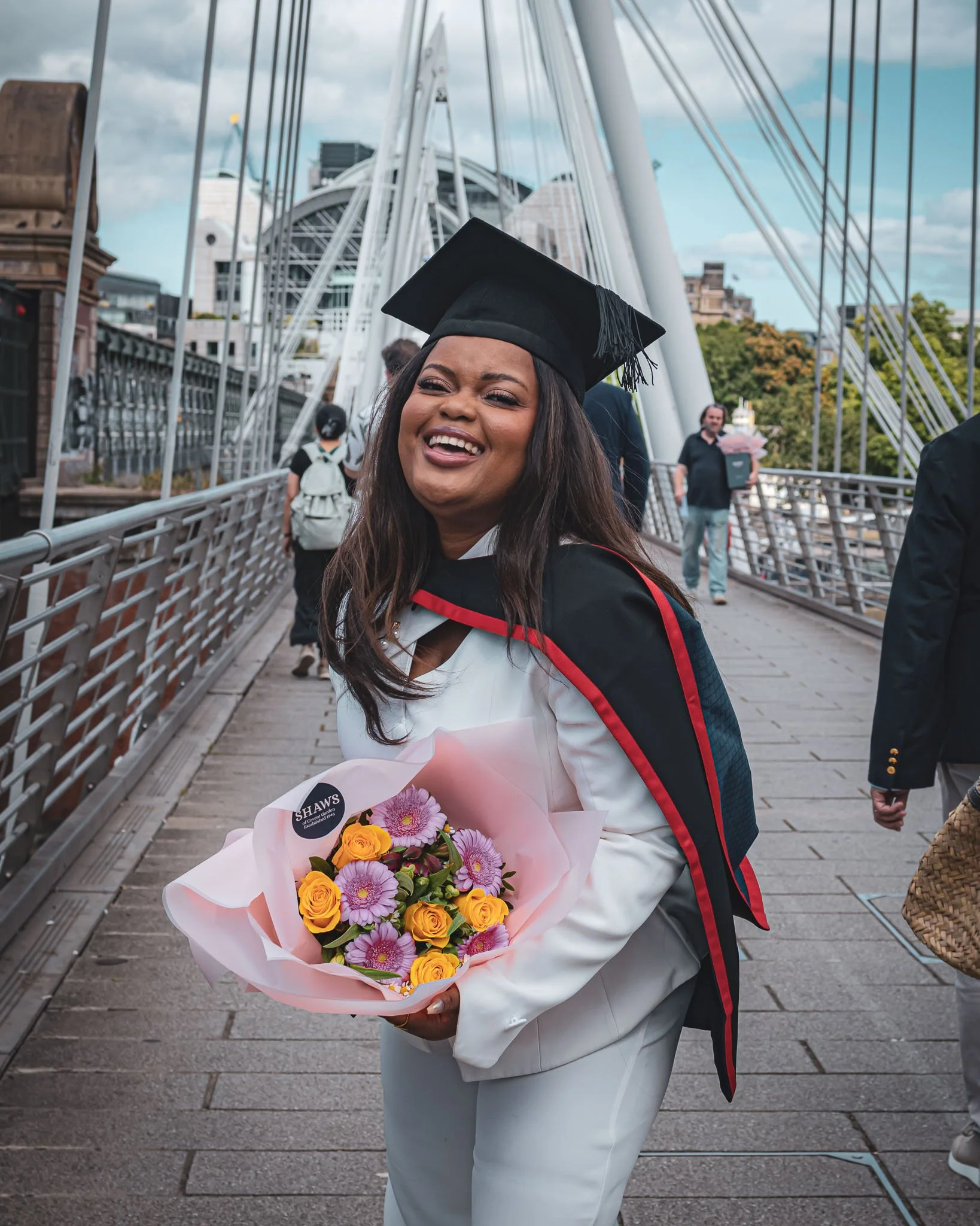 A woman in a graduation cap and gown holding a bouquet of flowers, smiling on a bridge with the cityscape in the background.