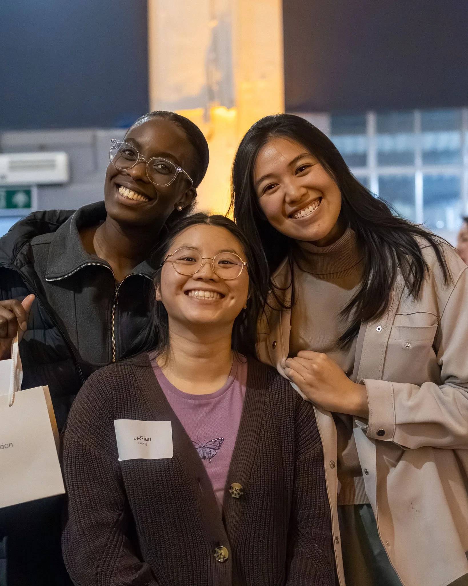 Three women smiling at an indoor event, one wearing glasses and a black jacket, another with a name tag, and another dressed casually.