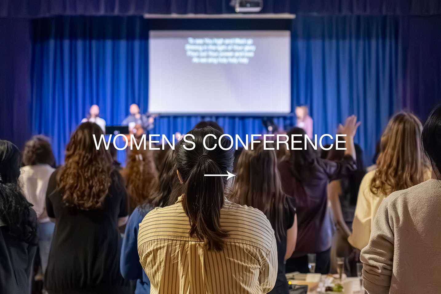 Women attending a conference in front of a stage with a screen, some raising their hands.
