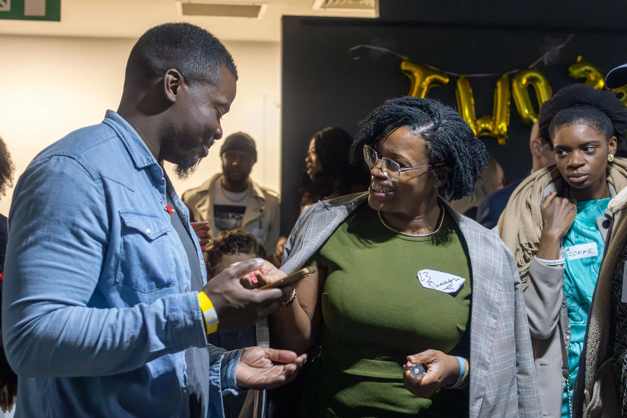 A diverse group of people gathered at an indoor event, engaging in conversations and interactions. Two women are prominently featured in the foreground, smiling and exchanging contact information. Decorated with gold balloons spelling out a word, pos