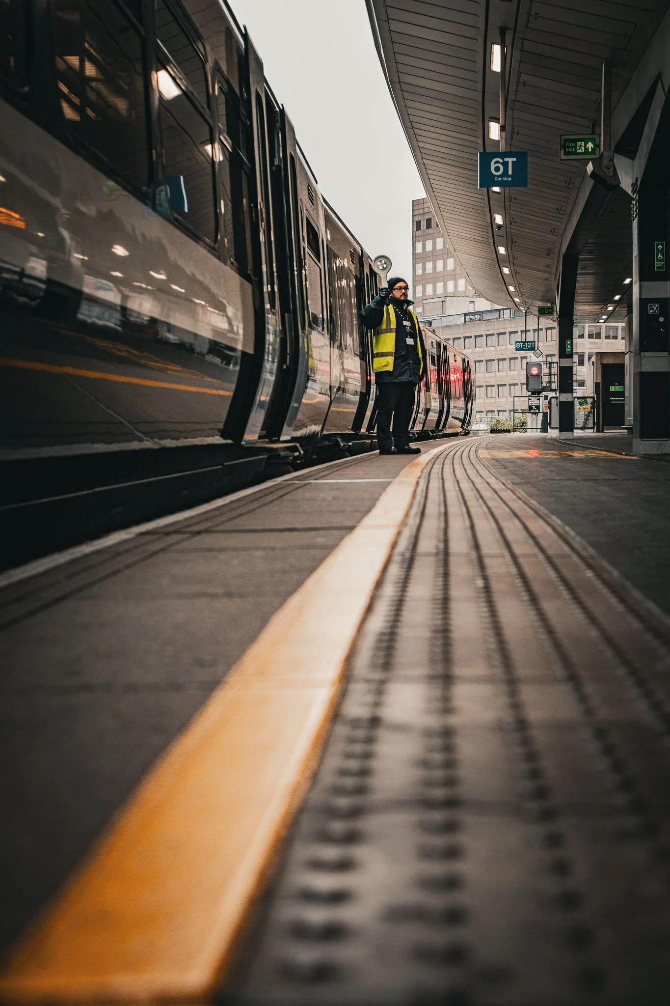 A person wearing a yellow safety vest and black gloves standing on a train platform at London Bridge holding a flashlight, with a train on the left side and a cityscape of tall buildings in the background.