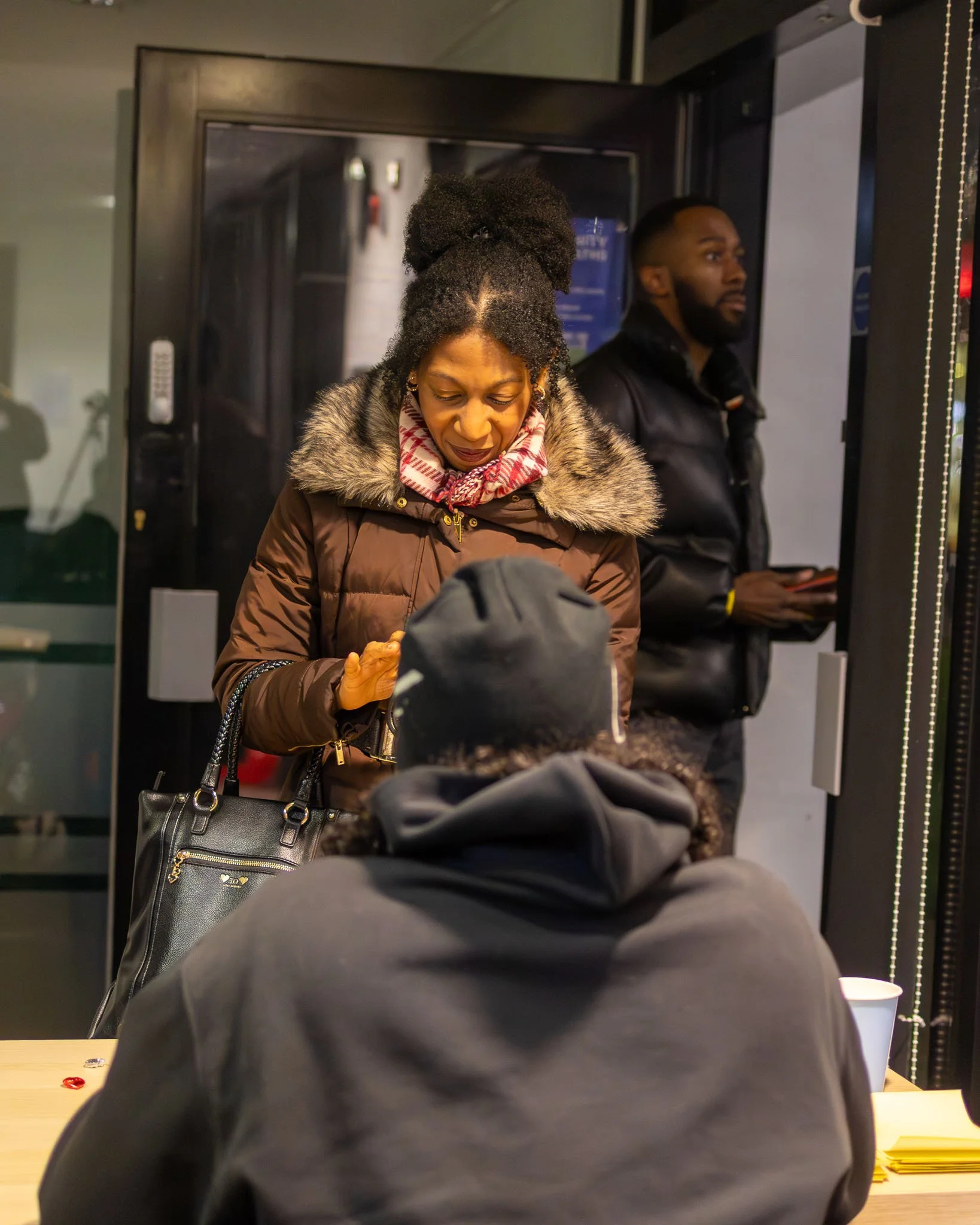 Two people standing and sitting inside a building near a vending machine. The woman in the foreground has her hair styled in a high bun and is wearing a fur-lined coat and scarf, looking down at her phone. The man in the background has a beard, is we