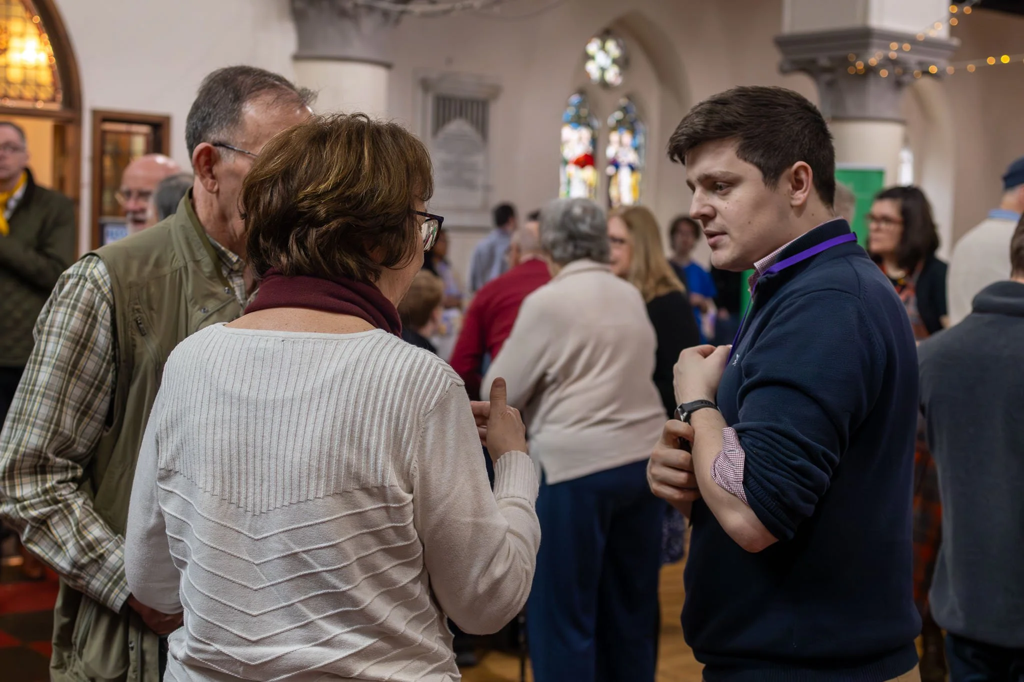 Two men and a woman having a conversation in a crowded indoor space, possibly a church or community hall, with stained glass windows and other people in the background.