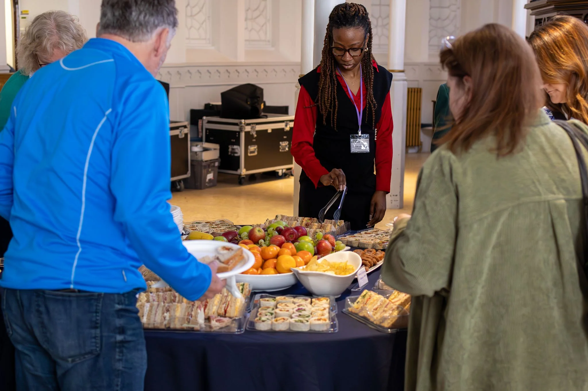 People serving themselves food at a buffet table filled with fruits, sandwiches, and snacks in a room with elegant decor.