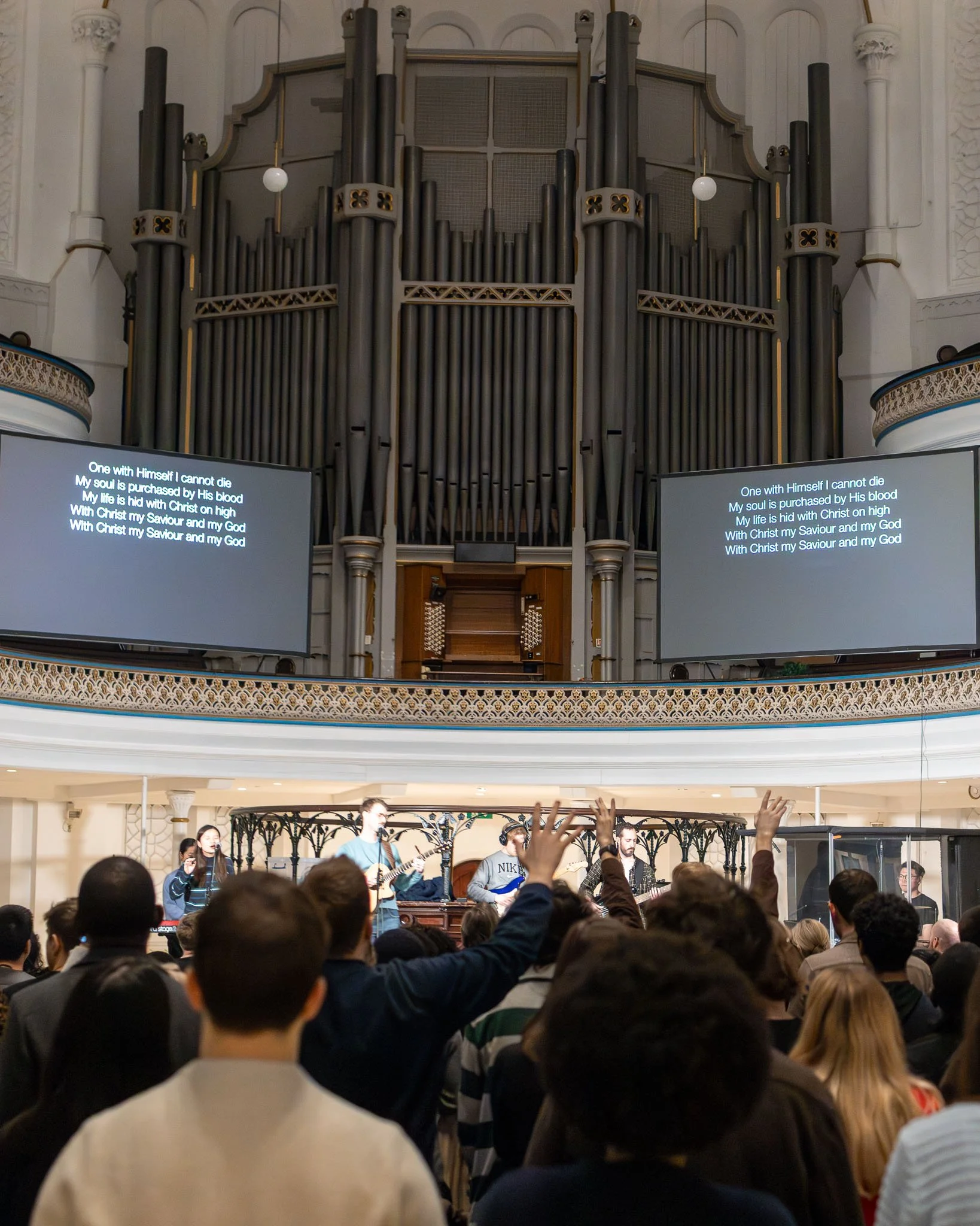 A worship service with a band and congregation in a church. The band features multiple musicians playing instruments and singing, with some congregation members raising their hands. Large screens display lyrics, and a grand pipe organ is visible in t