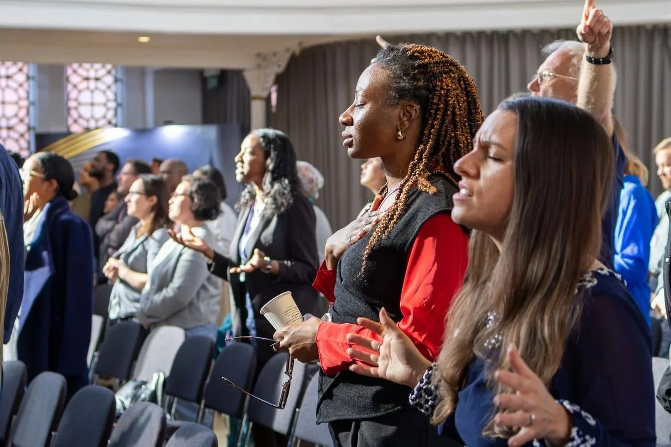 Group of diverse people attending a conference, some with eyes closed, hand over heart, and others with eyes closed in prayer or reflection, standing in a room with curtains and decorative windows.